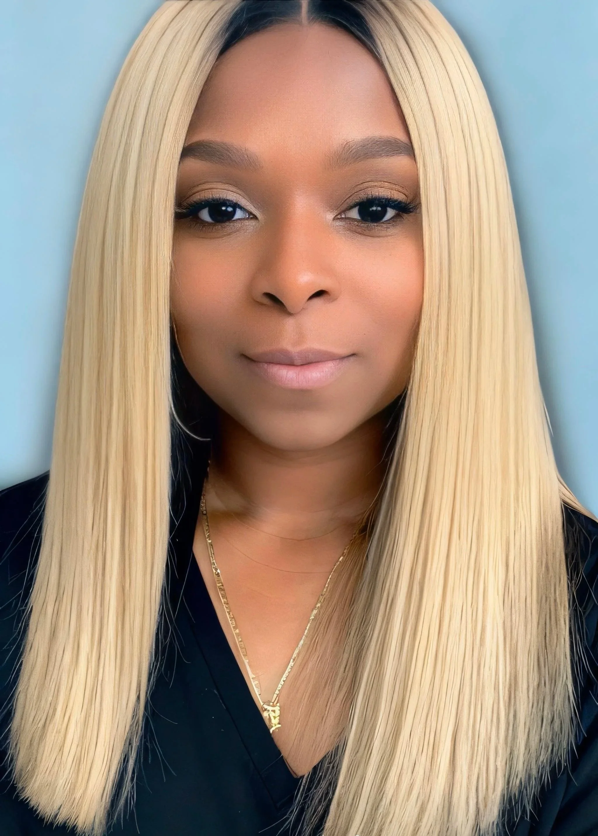 Close-up portrait of a woman with long straight blonde hair, wearing a black top and gold necklaces, against a plain light blue background.