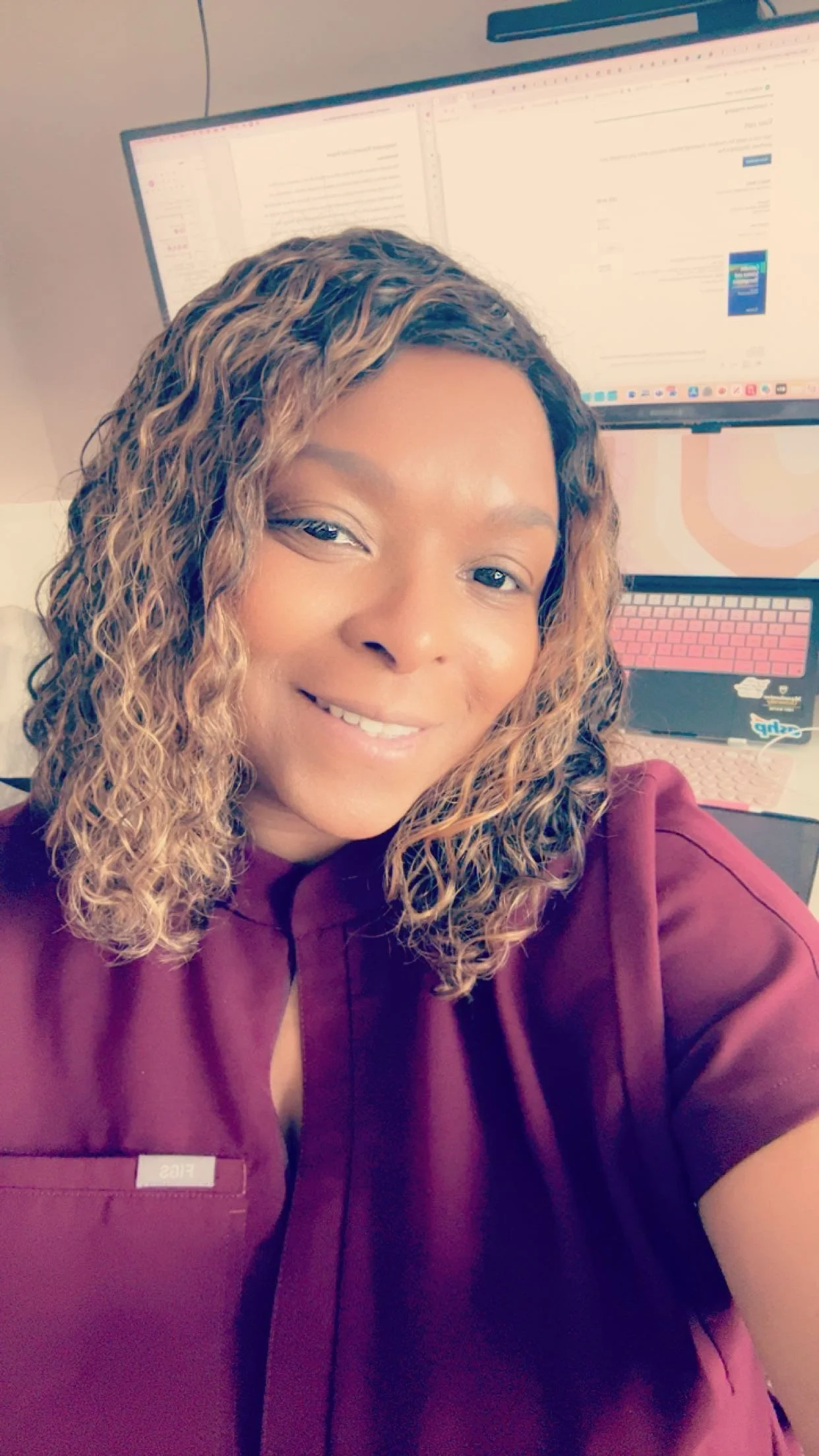A woman with curly hair smiling in an office, wearing a burgundy medical uniform, with dual computer monitors behind her showing a spreadsheet or document.