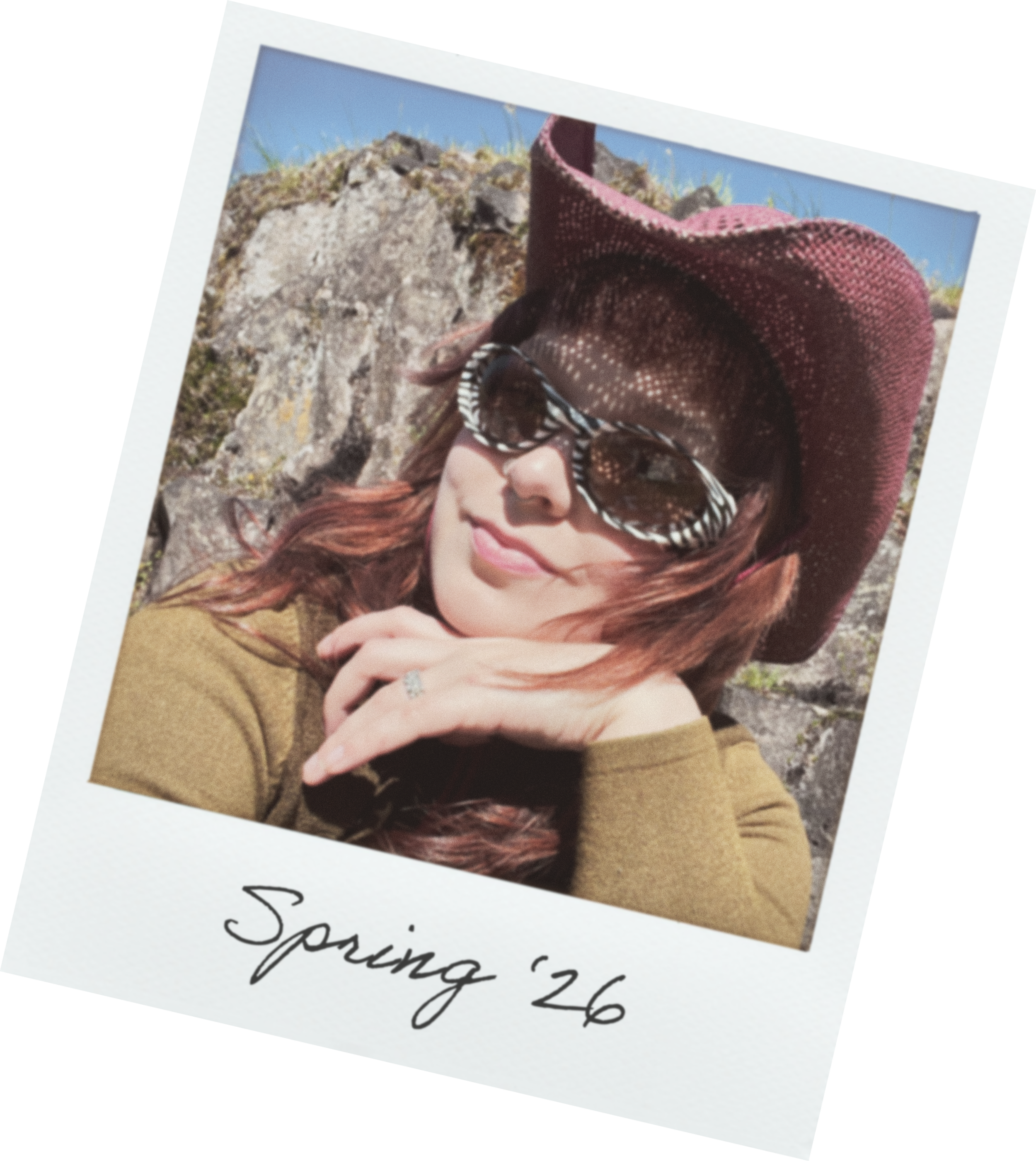A woman wearing a large burgundy sunhat and sunglasses, smiling outdoors with rocks and sky behind her. Part of a handwritten note is visible with the words 'Spring 26'.