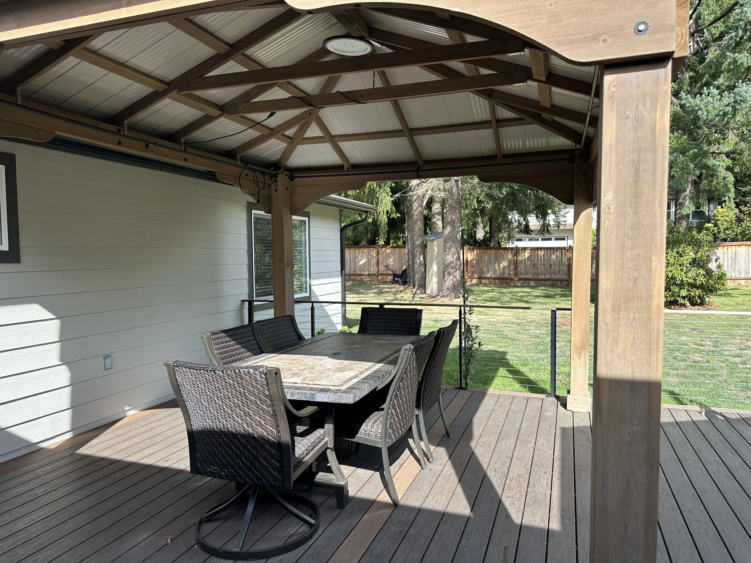 Outdoor patio with a dining table and six chairs under a wooden and metal roof, overlooking a backyard with trees and a wooden fence.