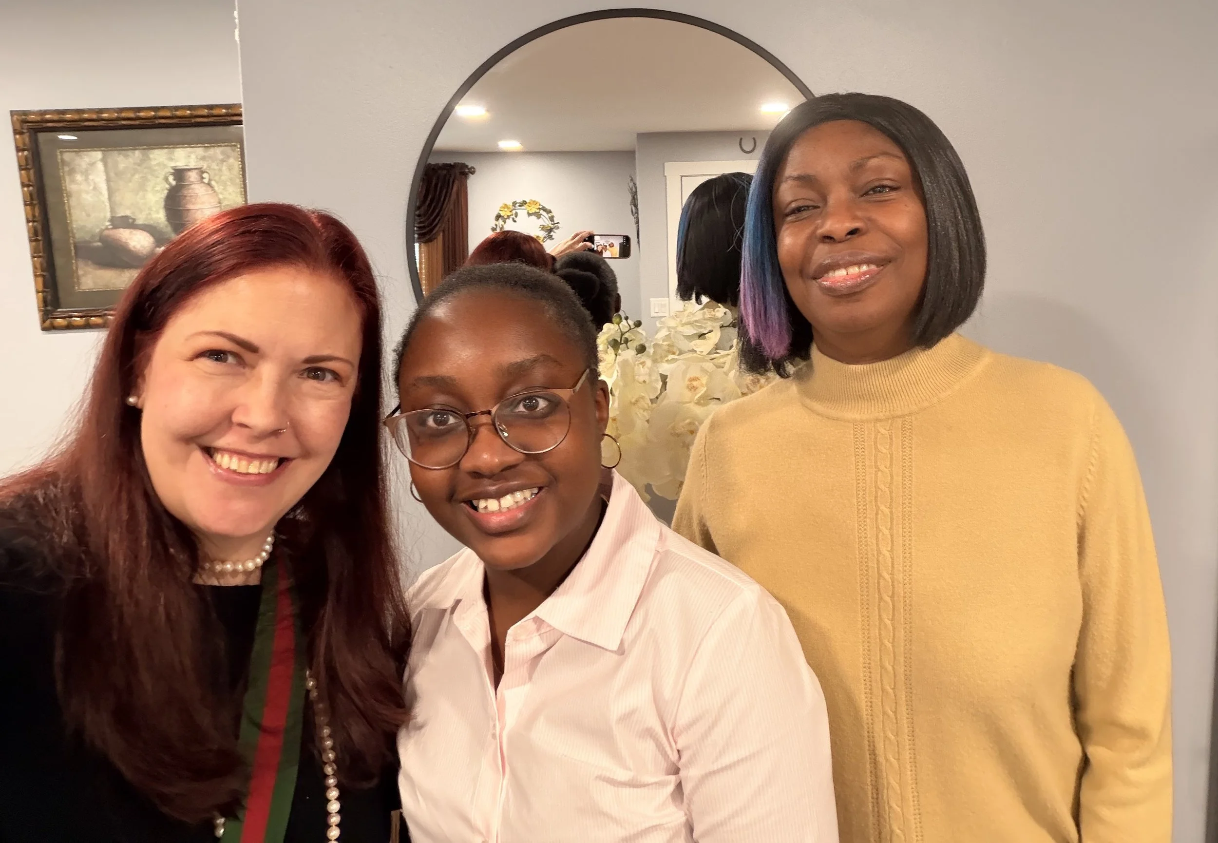Three women standing close together indoors, smiling at the camera. A woman with red hair wearing a pearl necklace and a black top, a young girl with glasses and a light pink shirt, and a woman with black hair with purple tips wearing a yellow sweater.