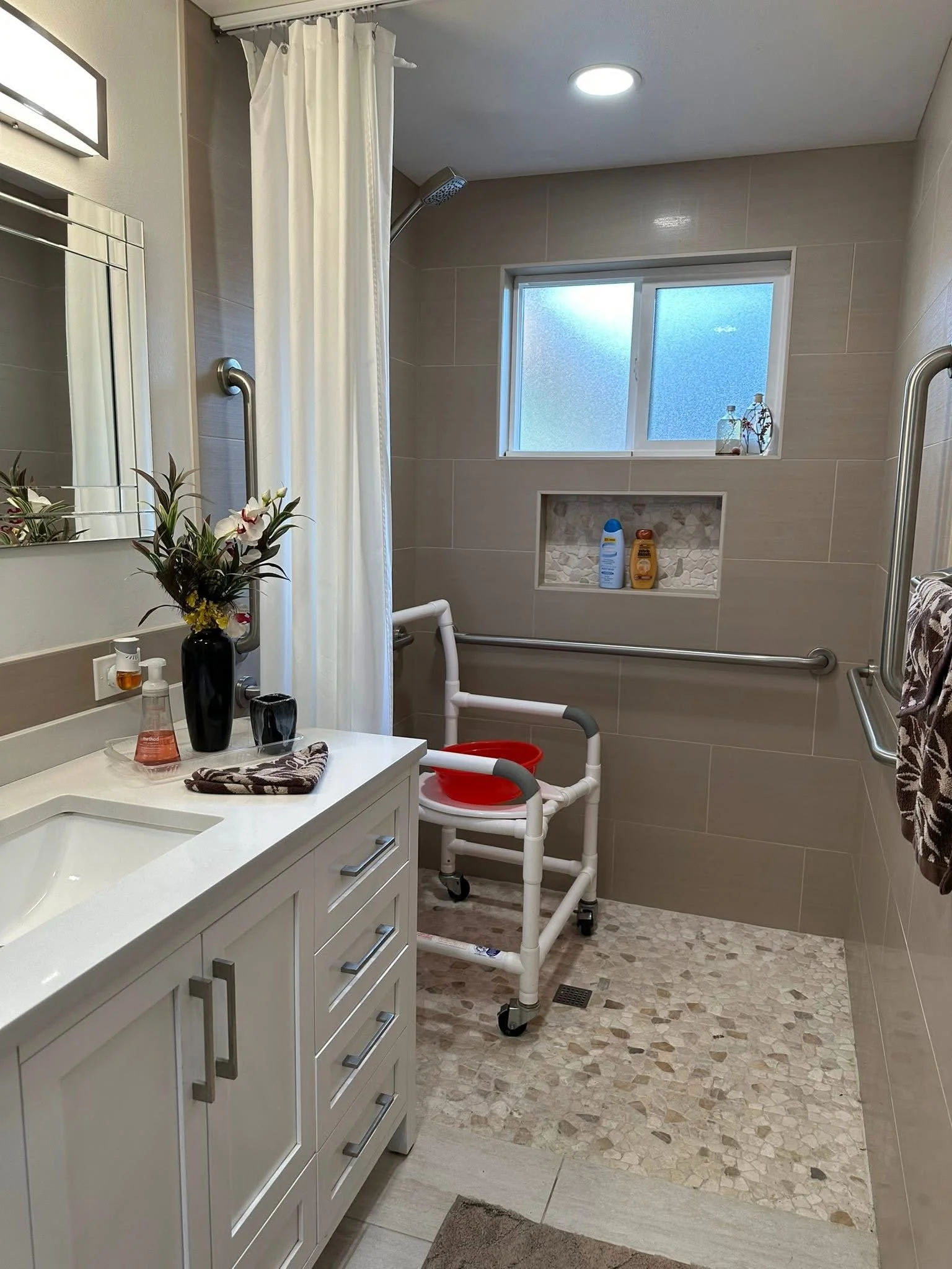 Bathroom with a white vanity, mirror, towels, soap dispenser, flower vase on the vanity, walk-in shower with a grab bar, a window, and a shower chair with a red basin.