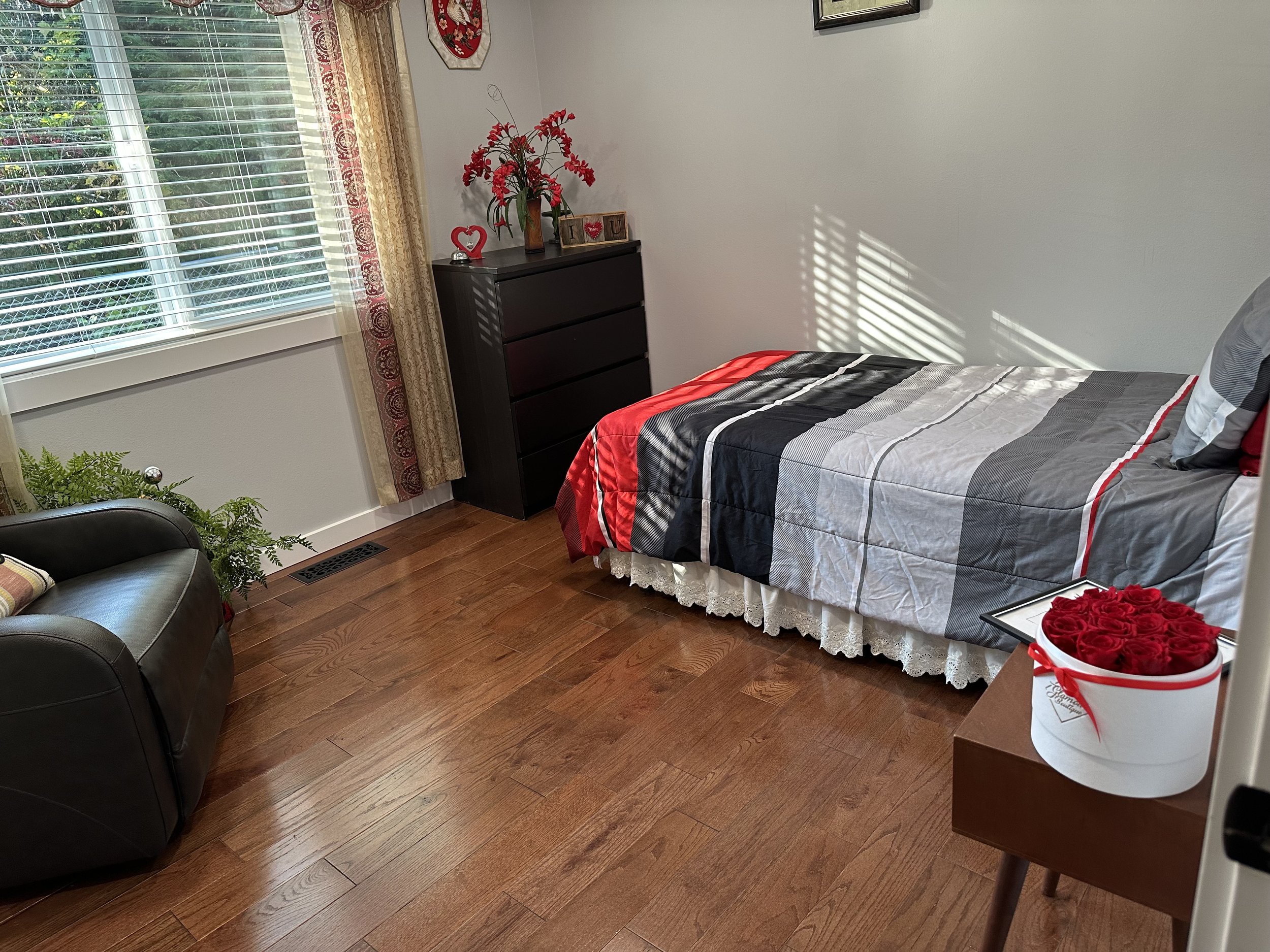 A bedroom with hardwood floors and a large window with blinds, decorated with flowers and framed pictures. There is a dark brown dresser with a vase of red flowers and ornaments, a bed with a colorful striped quilt, and a side table holding a white box of red roses.