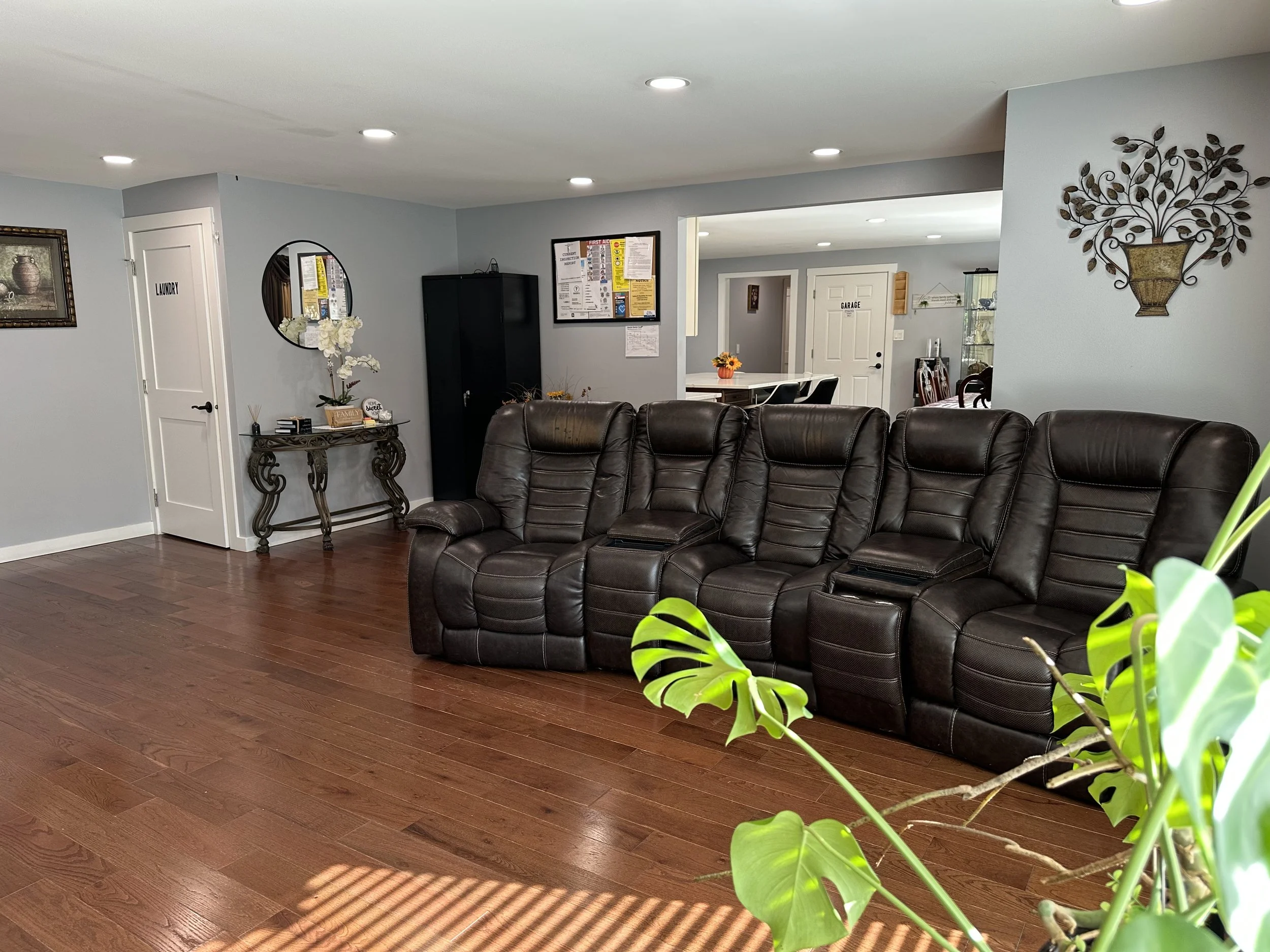 Living room with dark brown leather recliner sofa, wooden floors, light blue walls, decorative wall art, a side table with white flowers, a black cabinet, and a view into the dining area with a white door labeled 'Garage' and a table with a sunflower centerpiece.