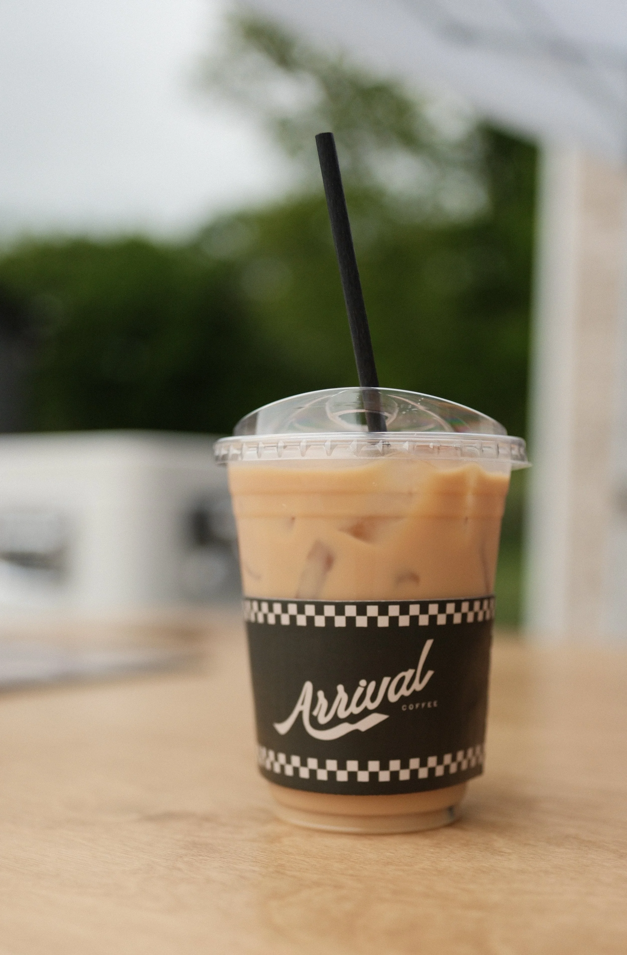 A to-go cup of iced coffee with a black straw, placed on a wooden table, with a blurred background of outdoor greenery. Coffee cart available for your next event. 