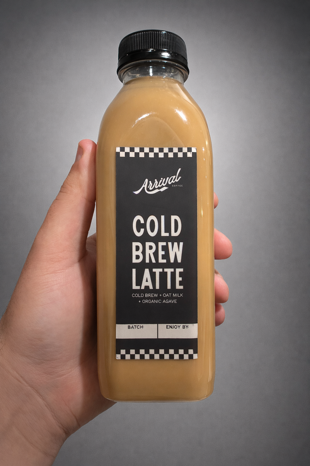 A hand holding a bottle of cold brew latte with a black cap and black label that reads "Arrival coffee, Cold Brew Latte, Cold Brew + Oat Milk + Organic Agave".