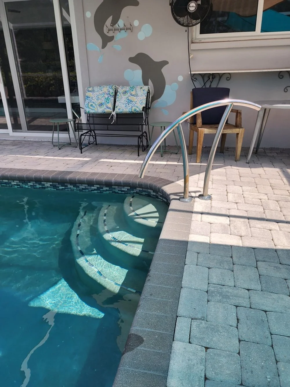 Backyard pool with steps, metal handrail, and brick border, featuring patio furniture including a bench with cushions, two small tables, and a wooden chair with a cushion, against a wall decorated with dolphin mural and a wall-mounted fan.