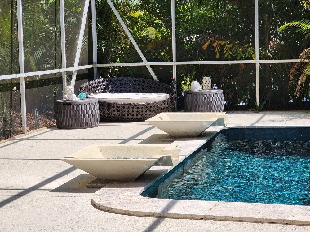 A backyard swimming pool with two white stone bowls on the deck, a black lounge chair, two wicker side tables, and lush green plants in the background.