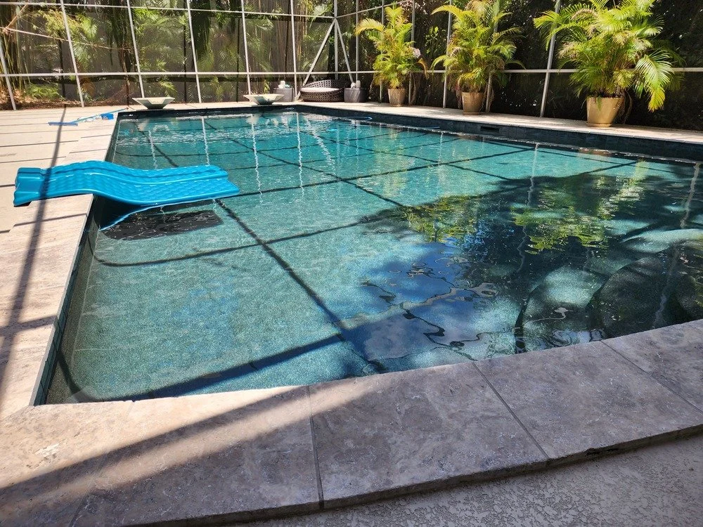 An indoor swimming pool with clear water, surrounded by a stone deck, potted plants along the back wall, and a blue swimming float in the water.