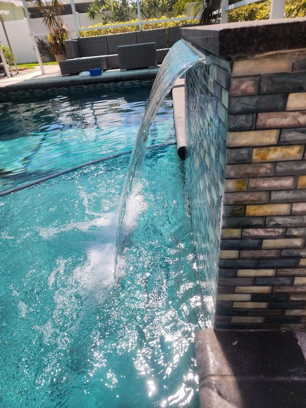 Close-up of a swimming pool water feature with water cascading from a tiled wall into the pool, patio furniture in the background, and sunny outdoor setting.