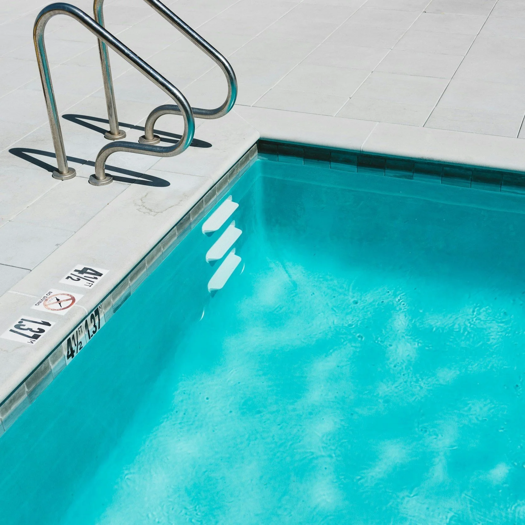 Empty swimming pool with metal handrails and white steps submerged in blue water, poolside tiled surface, and depth markers marked in black on white tile.