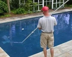 A man standing at the edge of a swimming pool, holding a pool skimming net, wearing a red cap, light t-shirt, and khaki shorts.