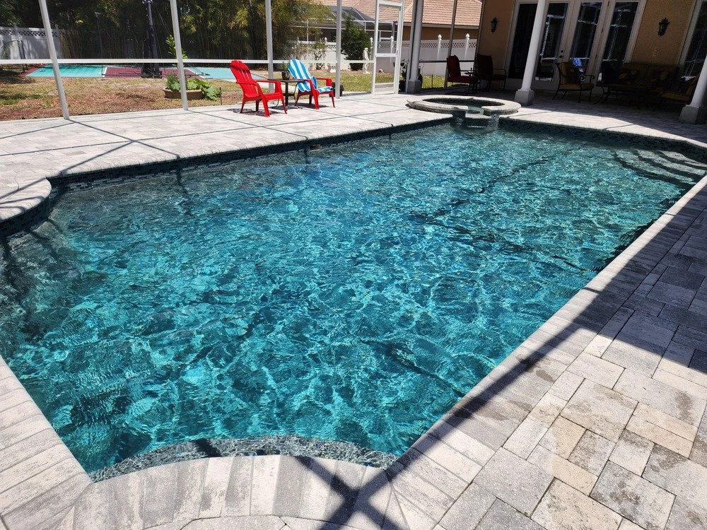 Backyard swimming pool with clear blue water, surrounded by paving stones, with pool chairs in the background.