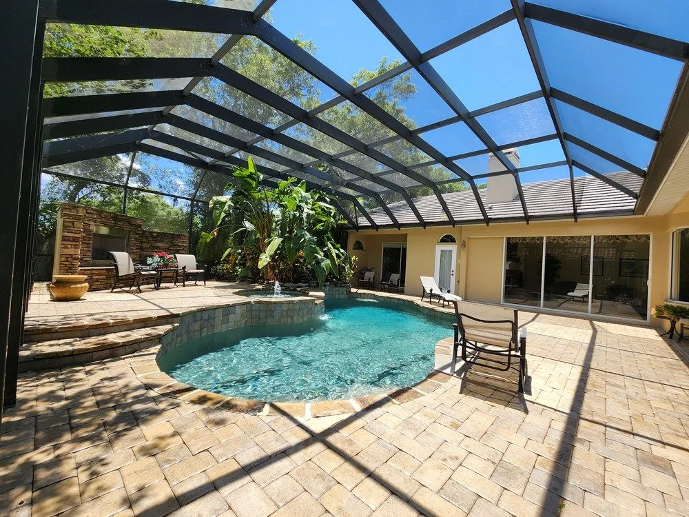 Enclosed backyard pool area with a screened-in patio, a small pool, outdoor seating, potted plants, and lush trees visible through the screen.