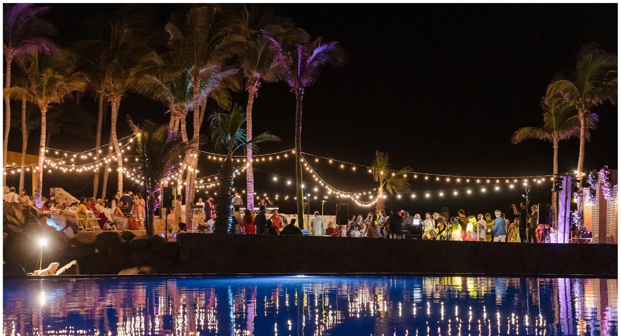 Nighttime outdoor party near a pool with string lights, palm trees, and groups of people celebrating.