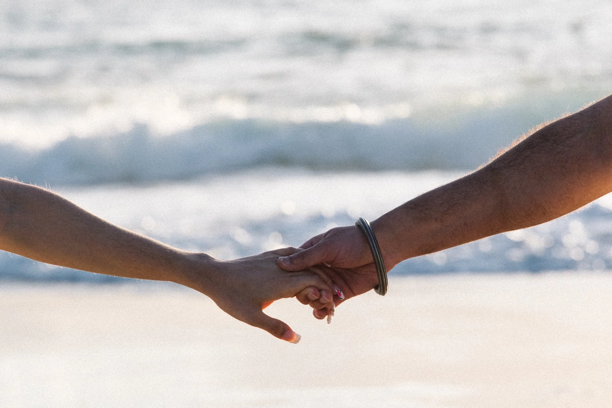 Two people holding hands on the beach, with ocean waves in the background.