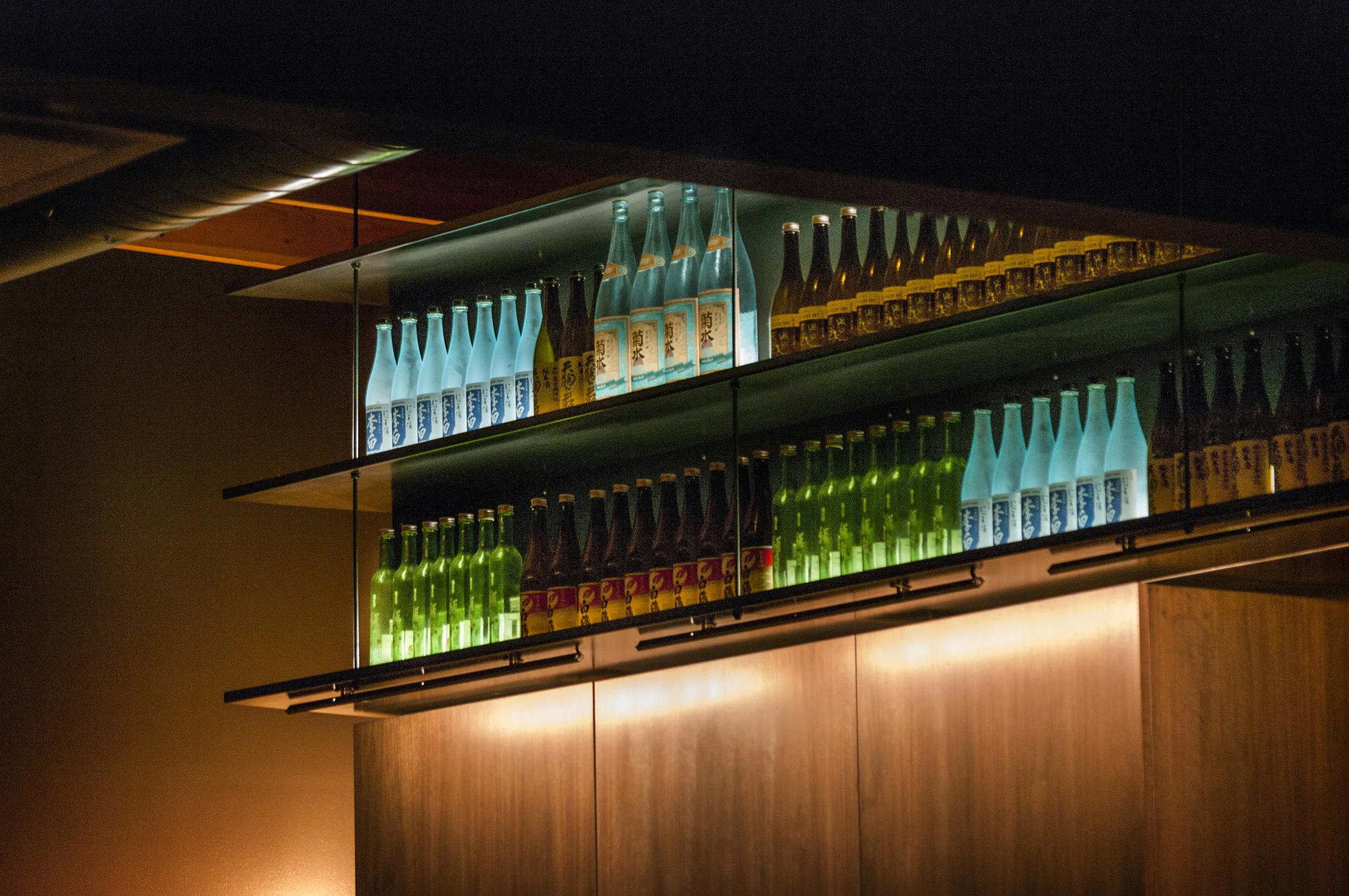 Illuminated glass bottle display with dark shelving and warm architectural lighting in a restaurant interior.
