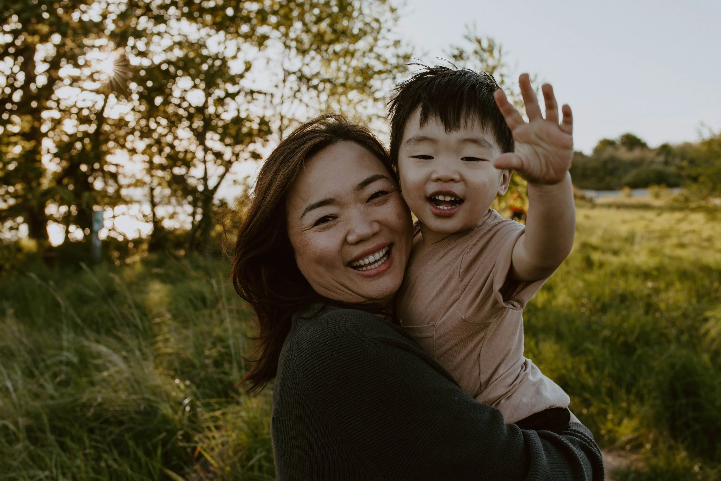 A woman with long dark hair and a young boy with short black hair are smiling happily together outdoors during sunset. The woman is holding the boy, who is reaching out with his hand, and they are surrounded by greenery with the sun shining through t
