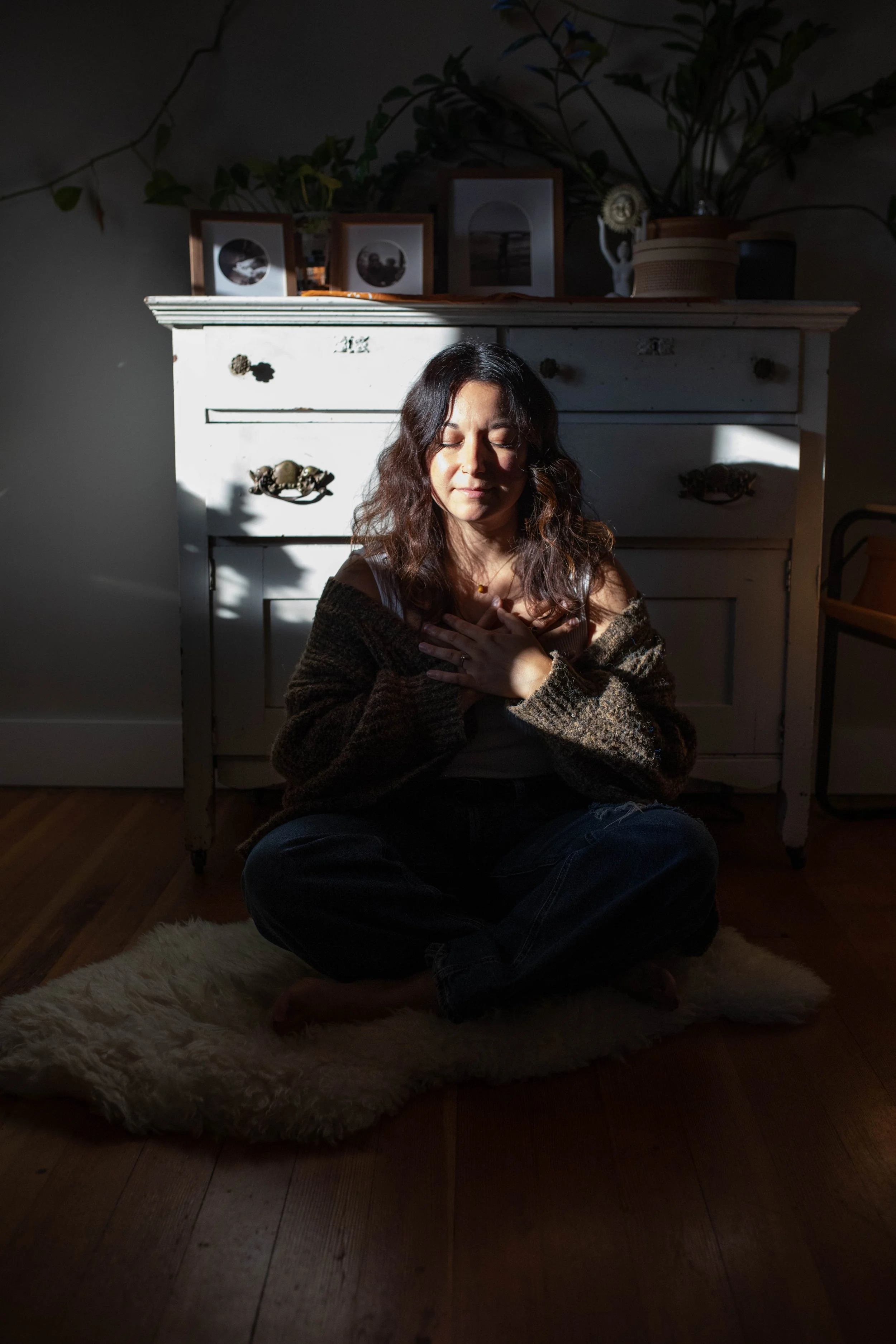 A woman sitting cross-legged on a fluffy rug with her eyes closed and hands on her chest, illuminated by a beam of sunlight in a cozy room with vintage white furniture and framed photos on top.
