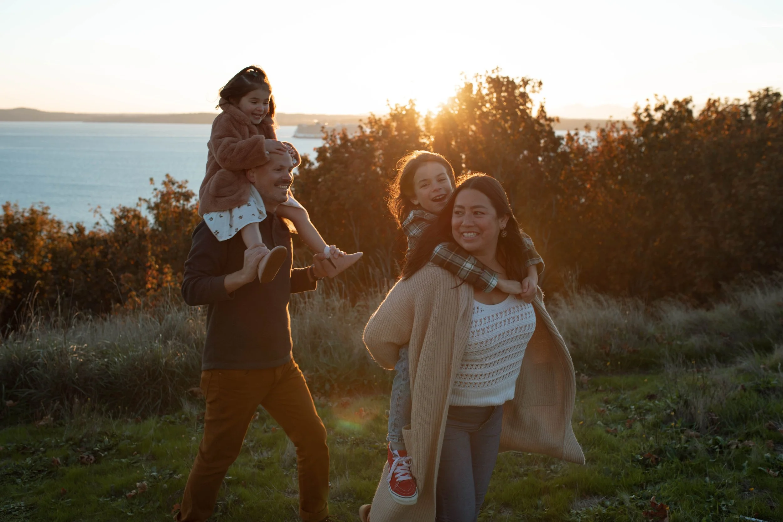 A family walking outdoors during sunset, with two parents and two children, by a body of water and trees.