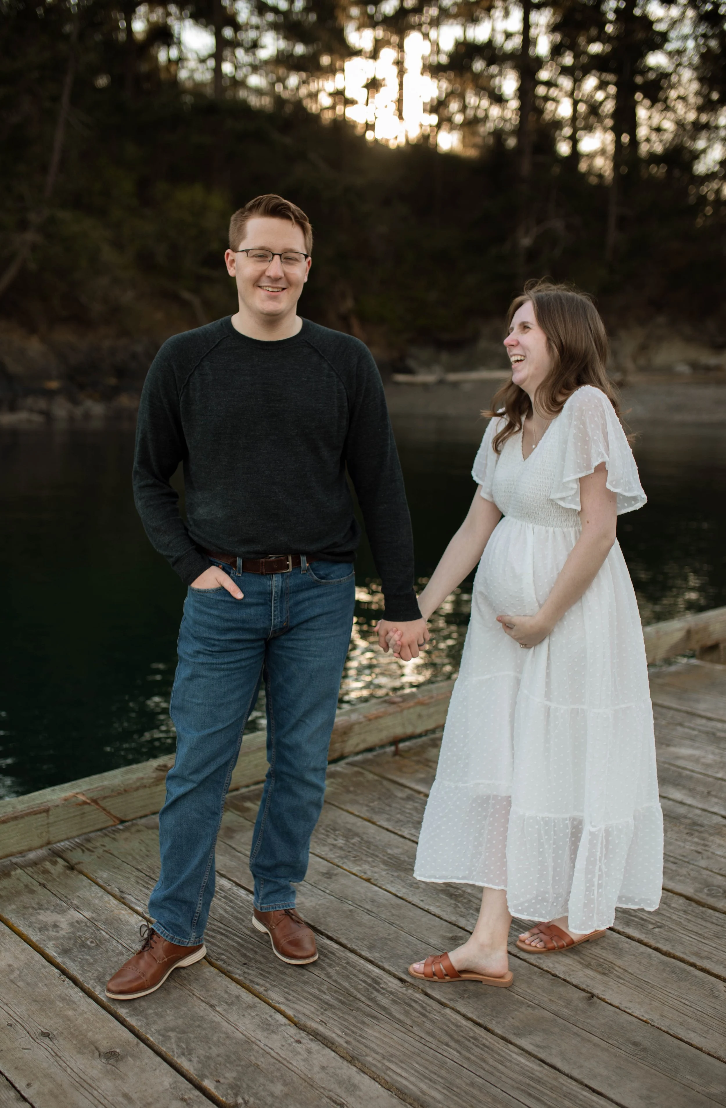 A happy couple stands on a wooden dock by a river, holding hands. The pregnant woman is smiling and gently touching her belly, while the man is smiling and looking at her. The background features trees and the setting sun.