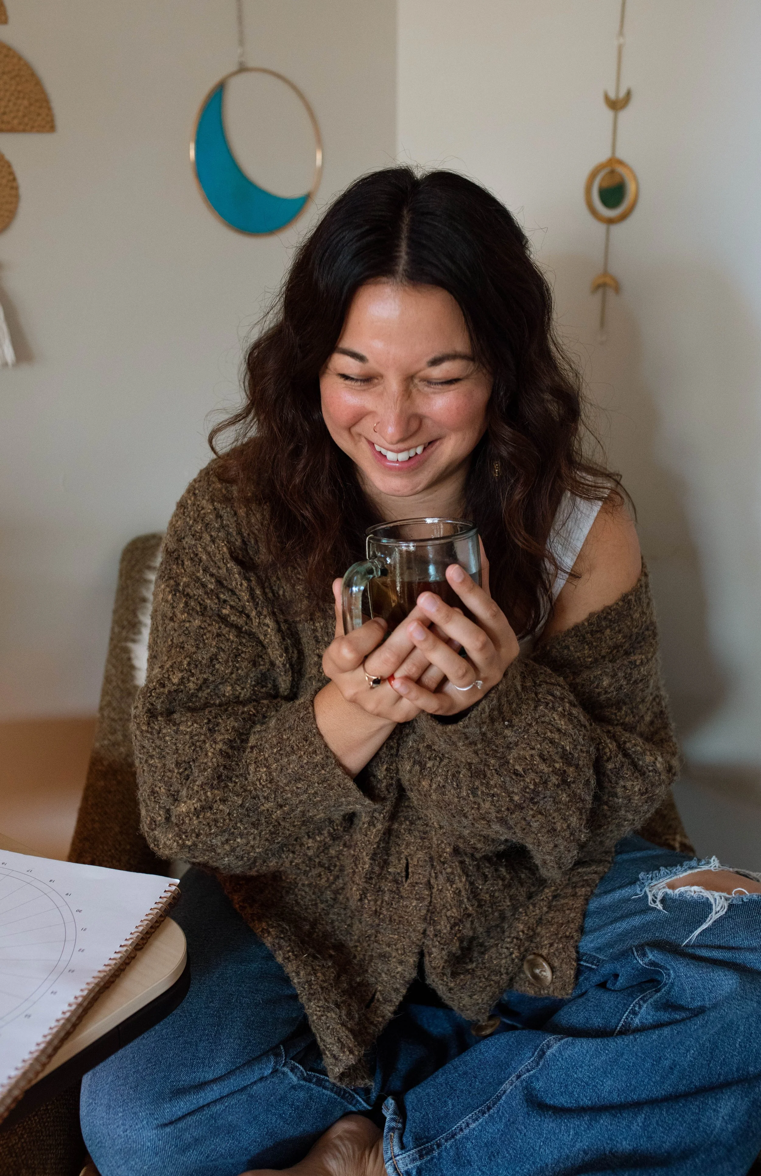 A woman with dark wavy hair, smiling, holds a cup of tea or coffee, sitting cross-legged in a cozy room with wall decorations.