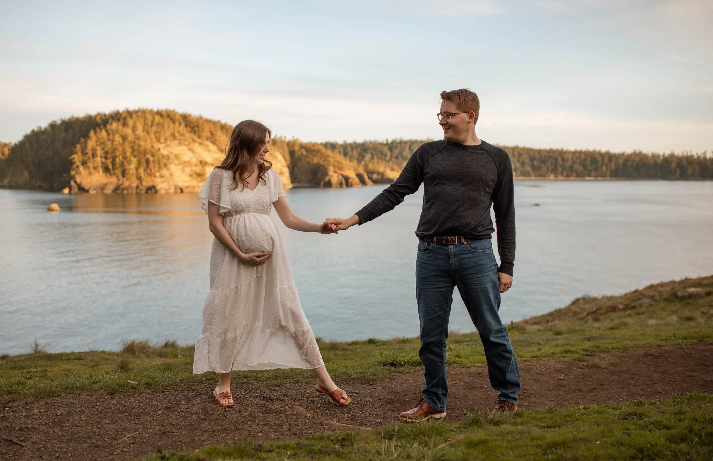 A pregnant woman wearing a white dress and sandals holding her belly with her left hand, stands on a grassy area near a body of water. A man in a black shirt, jeans, and glasses holds her right hand, smiling at her. The background features a scenic v