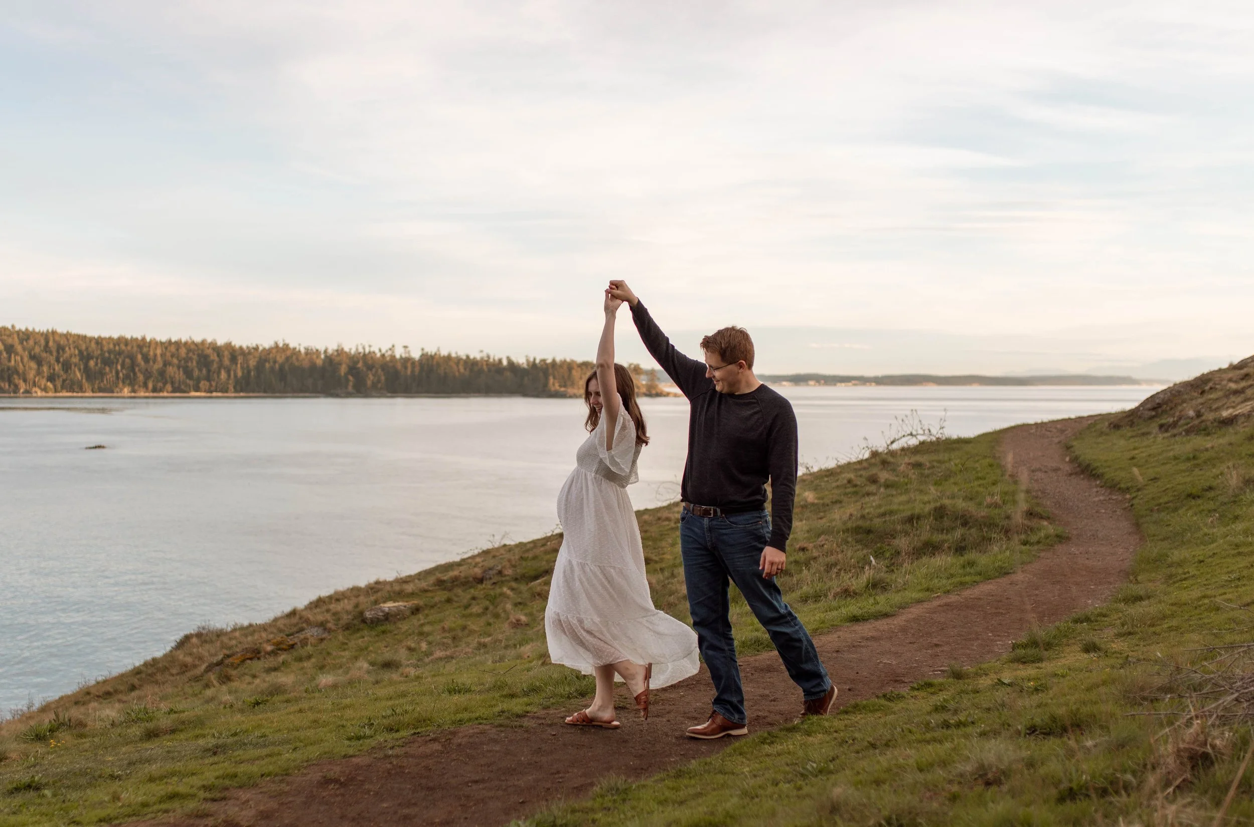 A couple dancing on a dirt path beside a river, holding hands with smiles, with a forest and calm water in the background.