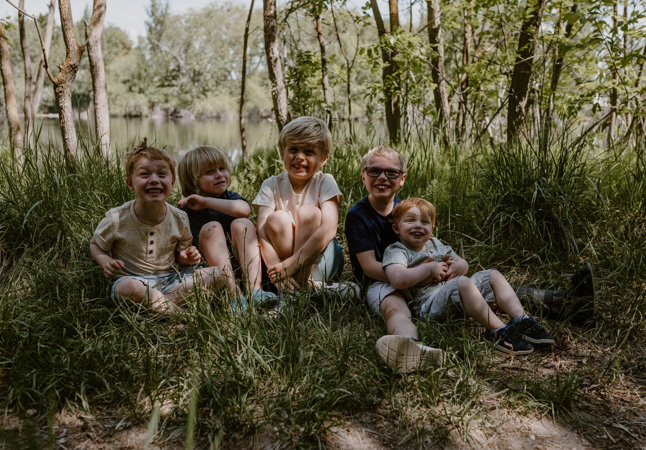 Five happy children sitting on the grass near a lake in a wooded area, smiling and enjoying the outdoors.