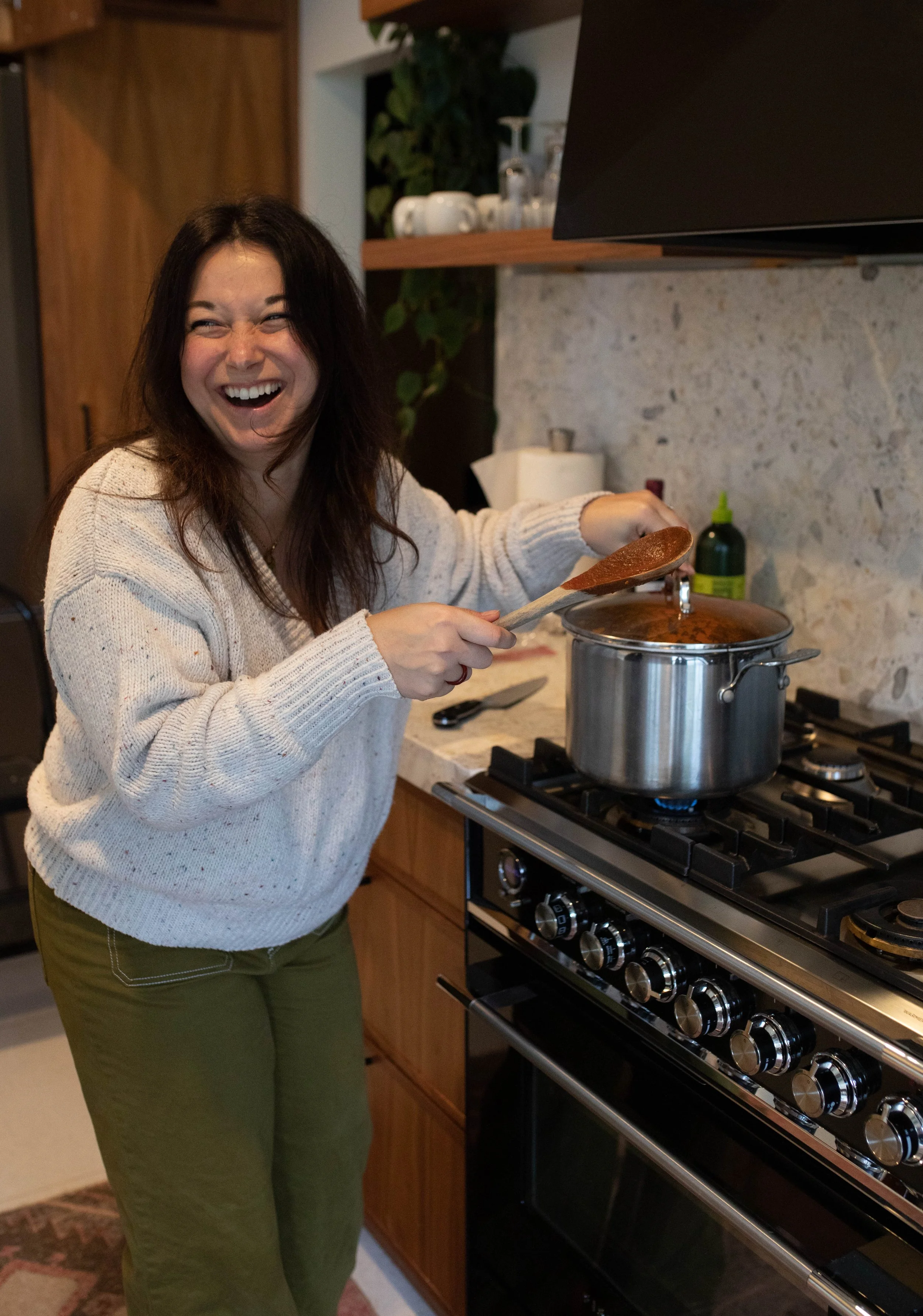 A woman wearing a cream sweater and green pants is cooking on a stove, stirring a pot with a wooden spoon while smiling and laughing.