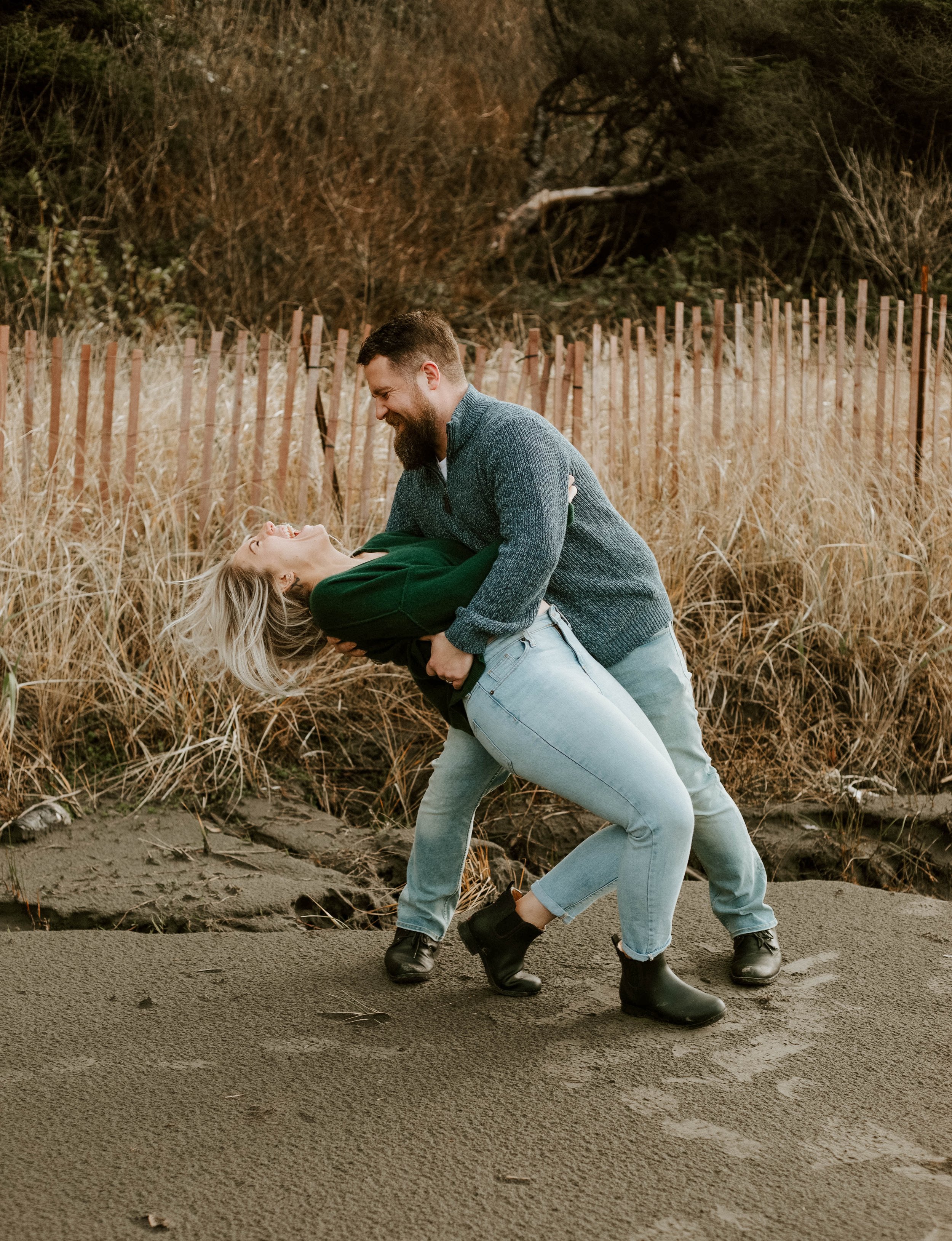 A couple dancing and smiling outdoors on a sandy path with tall dry grass and a wooden fence in the background.