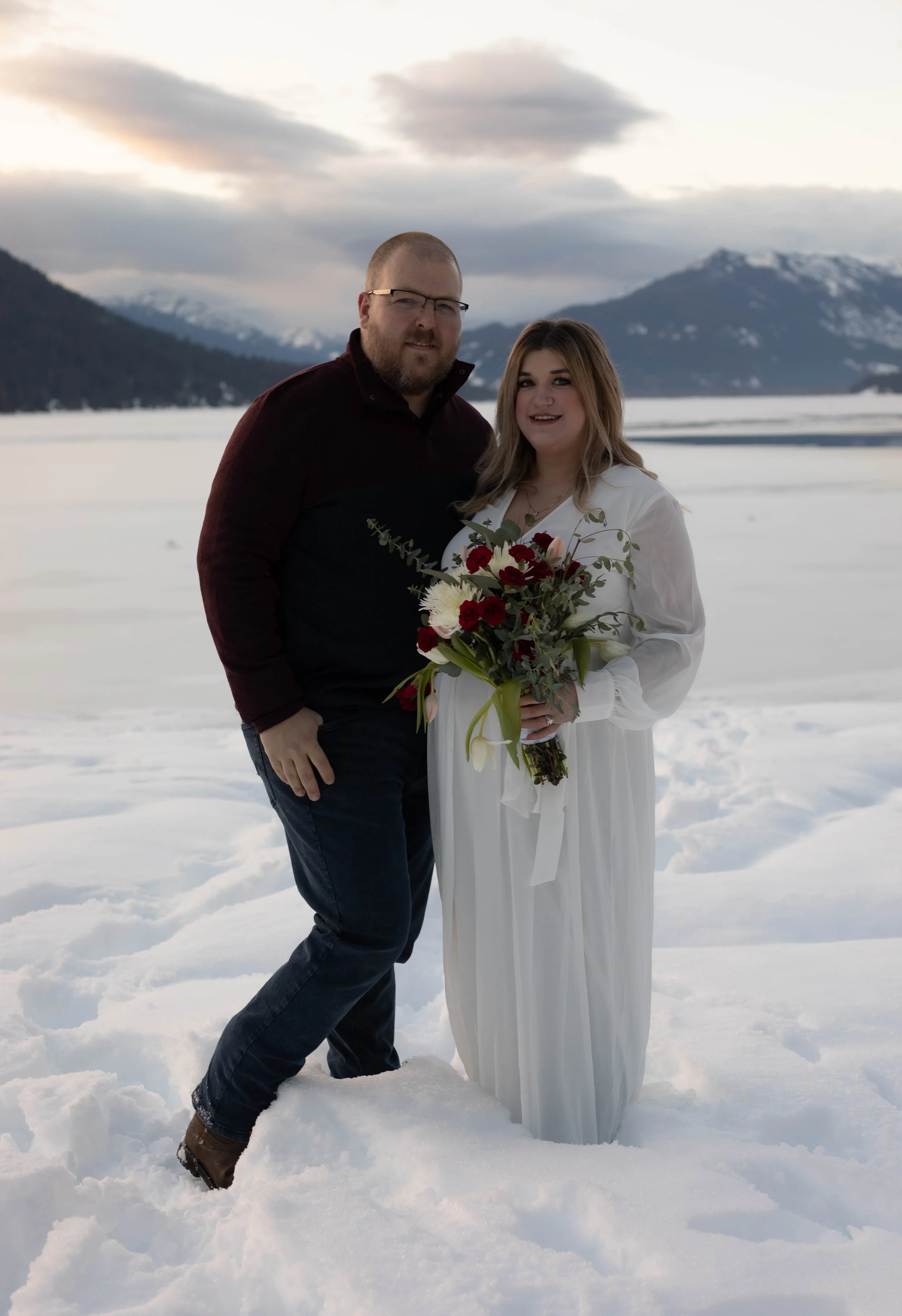 A couple standing in snow with snowy mountains and cloudy sky in the background; the woman is holding a bouquet of flowers.