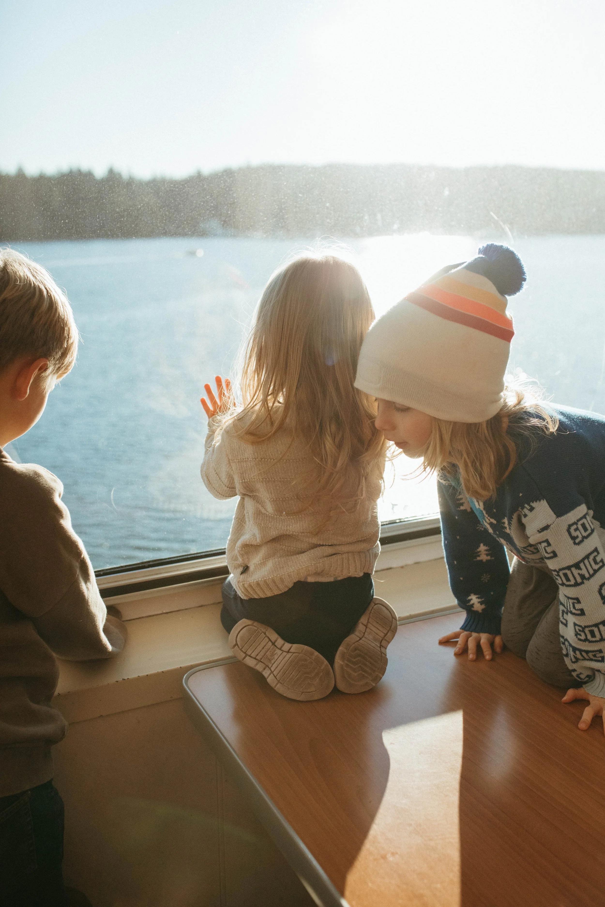Three children looking out a large window at a snowy landscape and lake outside on a sunny day.