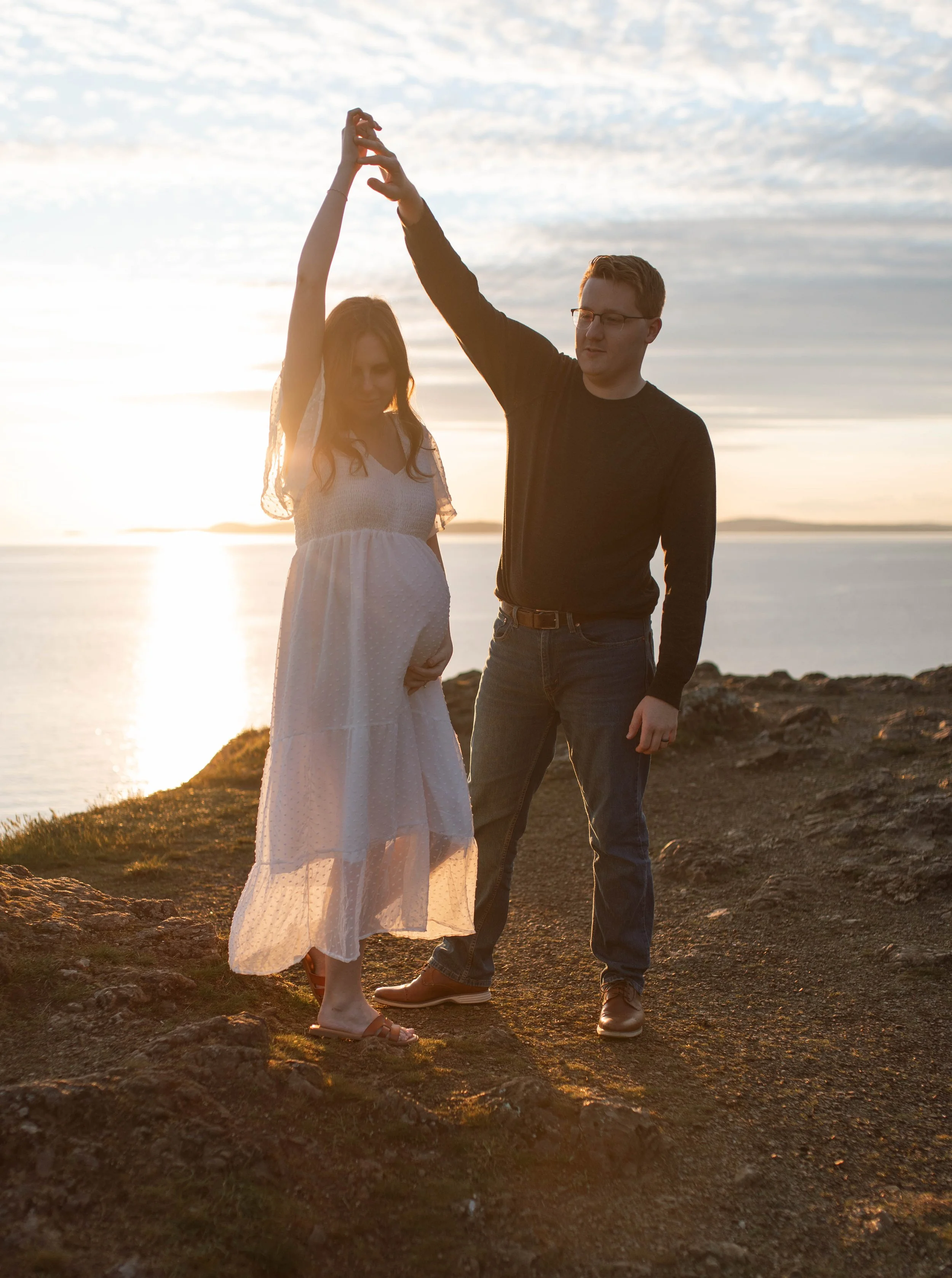 A couple standing on a rocky shoreline during sunset, with the woman pregnant, holding hands and touching the sky together.