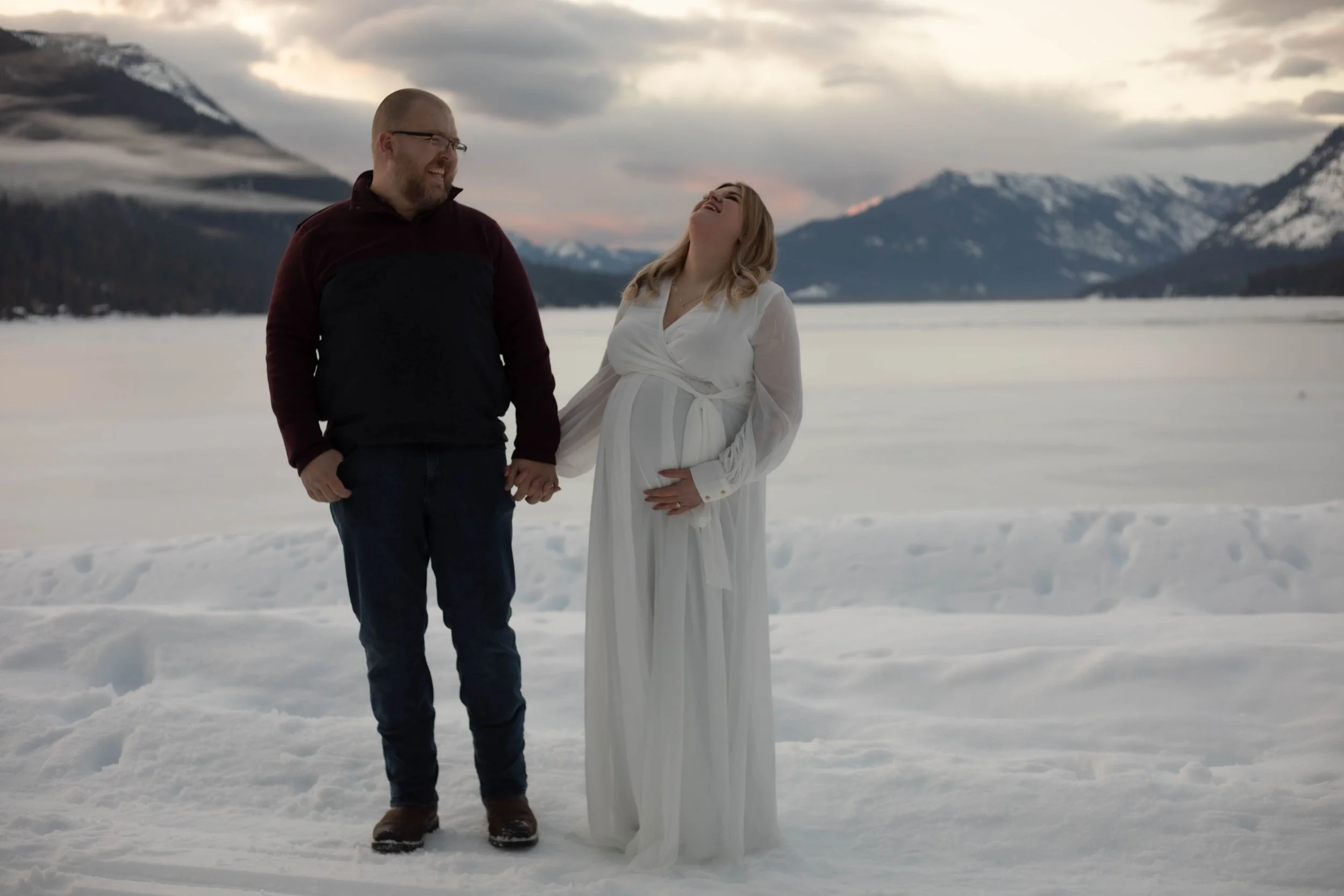 A couple standing in snow-covered landscape holding hands, with mountains and cloudy sky in the background during sunset.