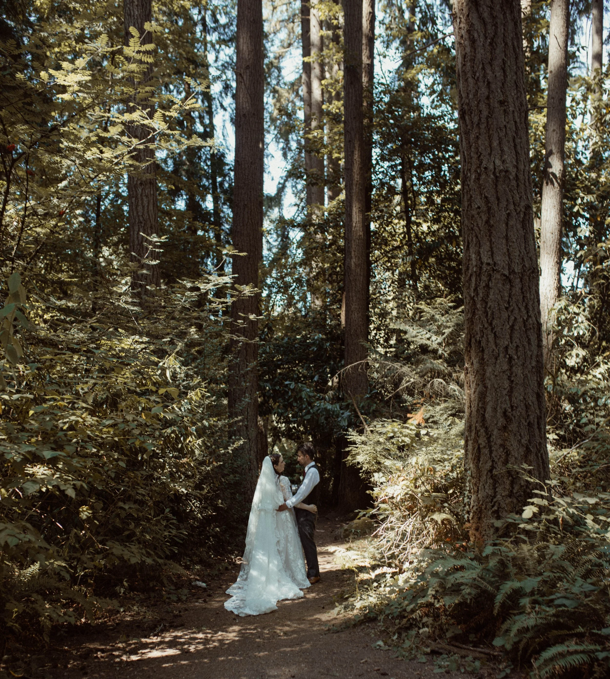 A bride and groom holding hands in a forest with tall trees and sunlight filtering through the leaves.