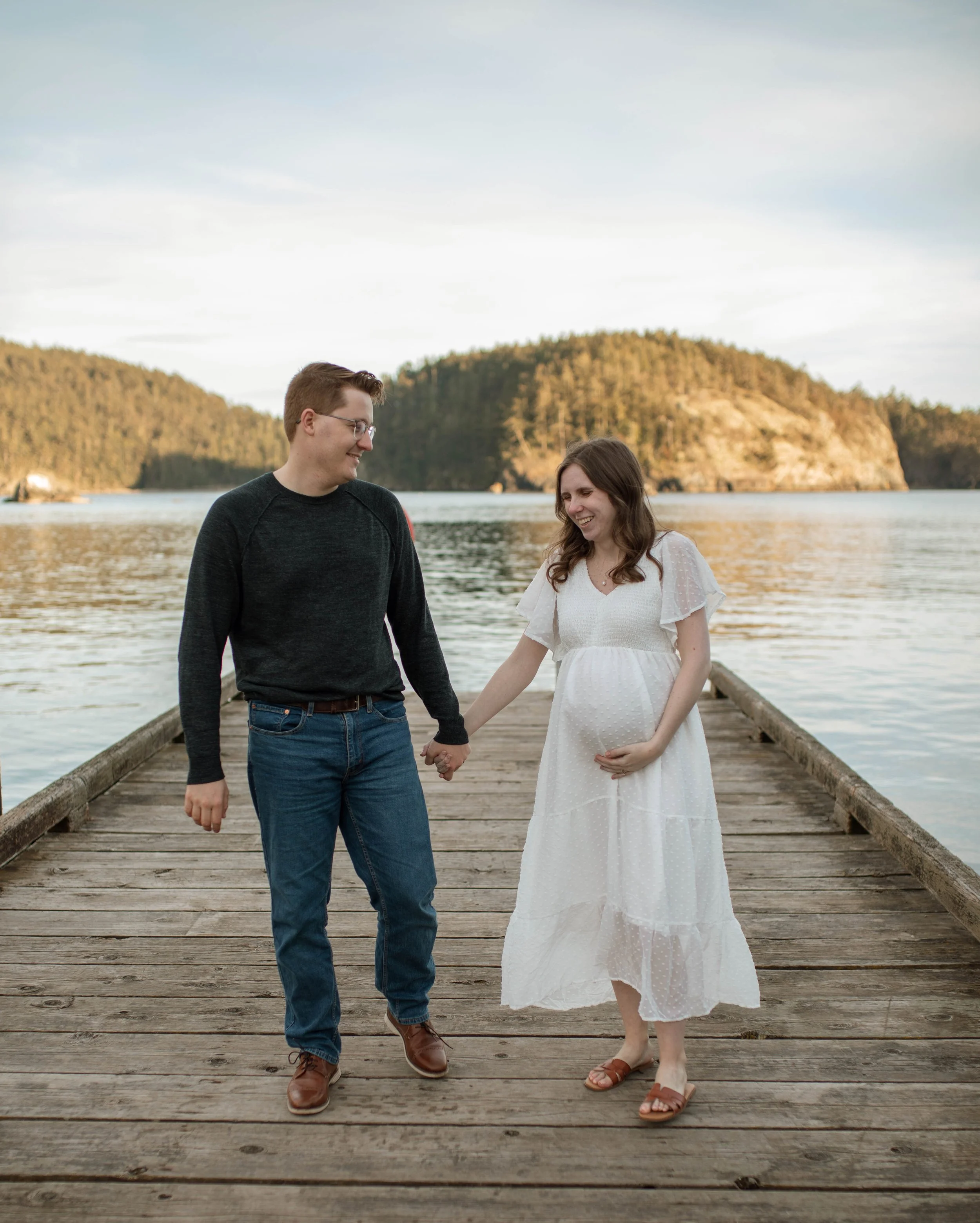 A pregnant woman and a man holding hands on a wooden dock by the water, with trees and hills in the background.