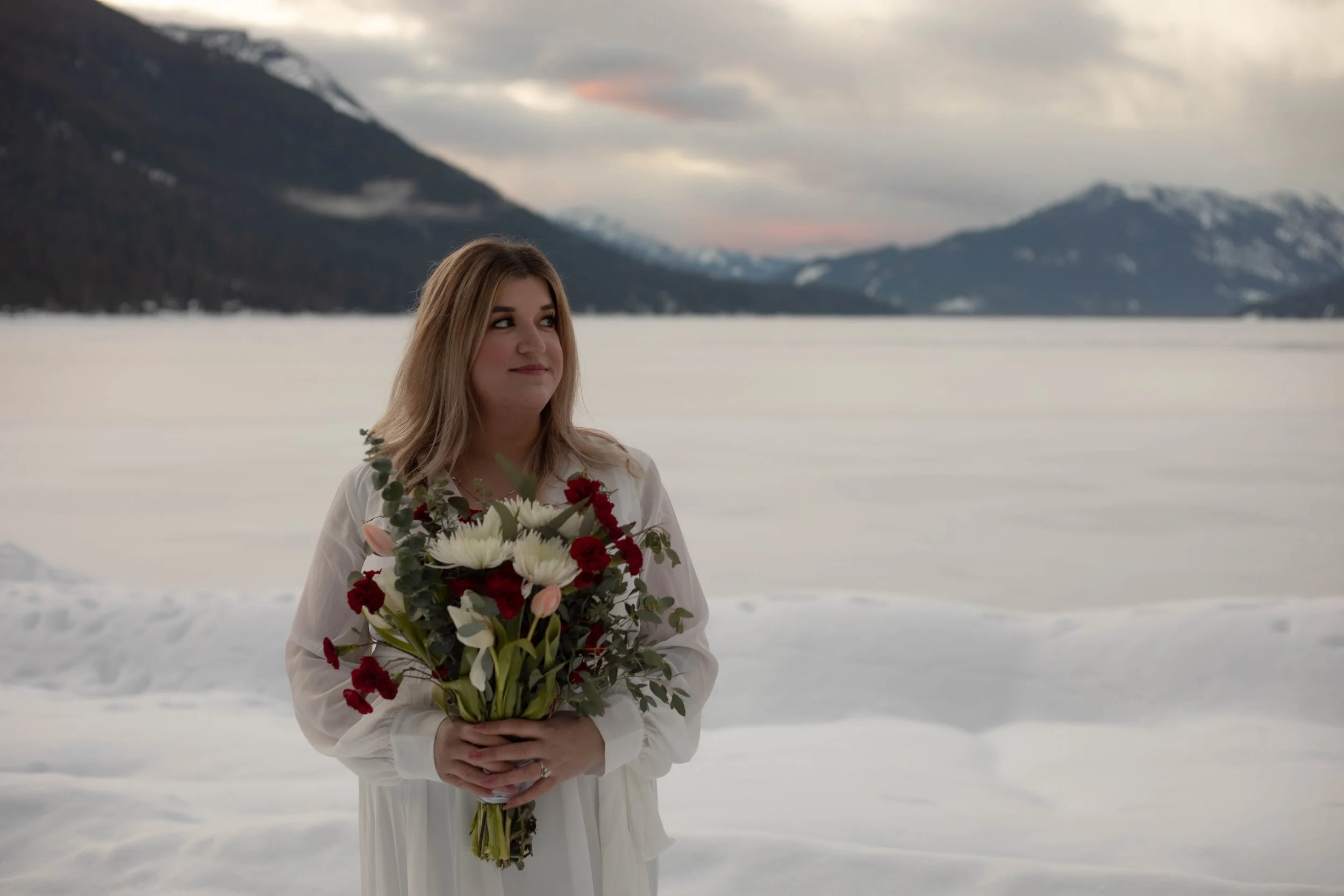 A woman with light brown hair, wearing a white dress, holding a bouquet of red, white, and pink flowers, standing in a snowy landscape with mountains in the background and a cloudy sky.