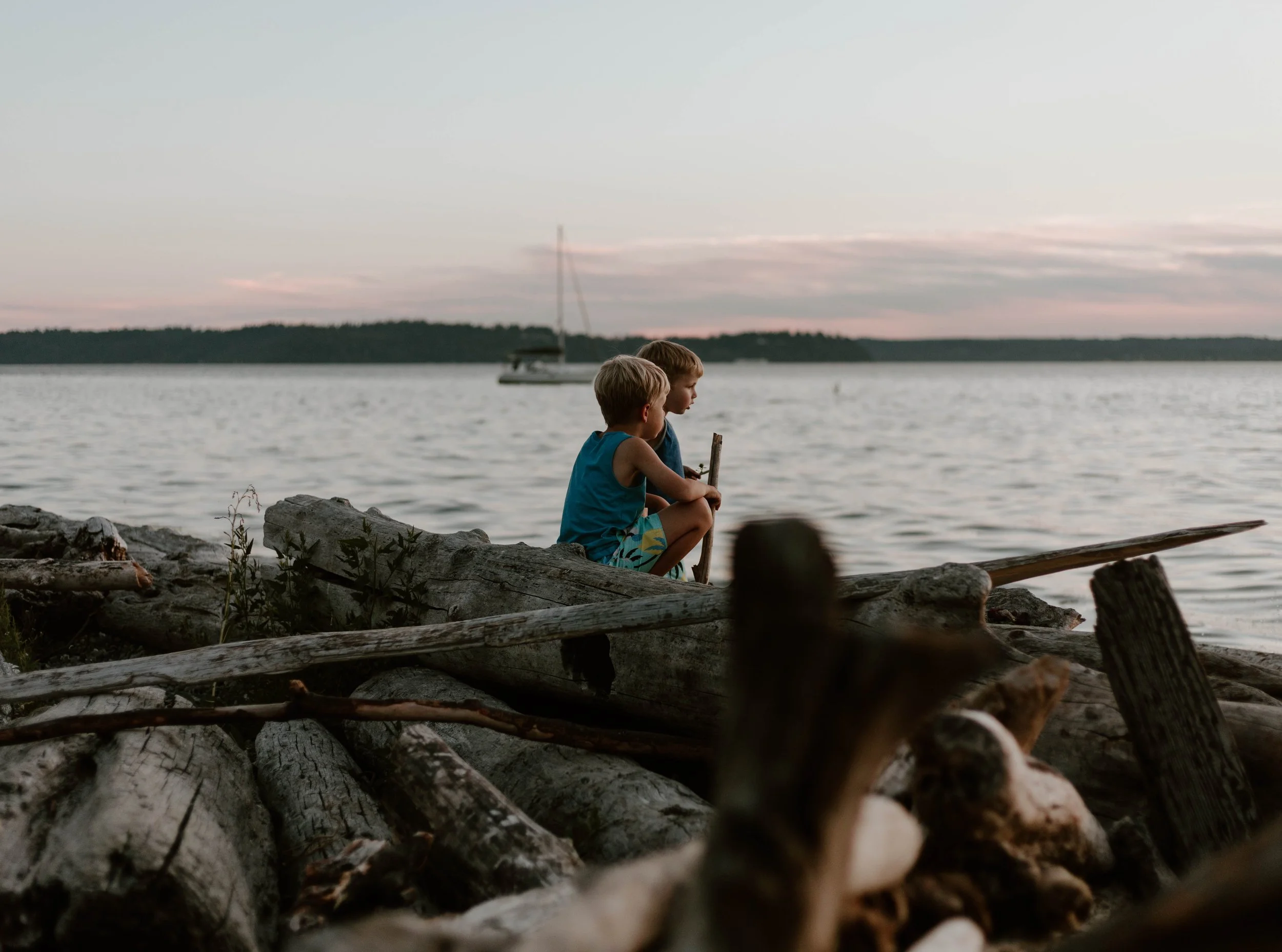 Two children sitting on driftwood near the water, watching a sailboat during sunset.