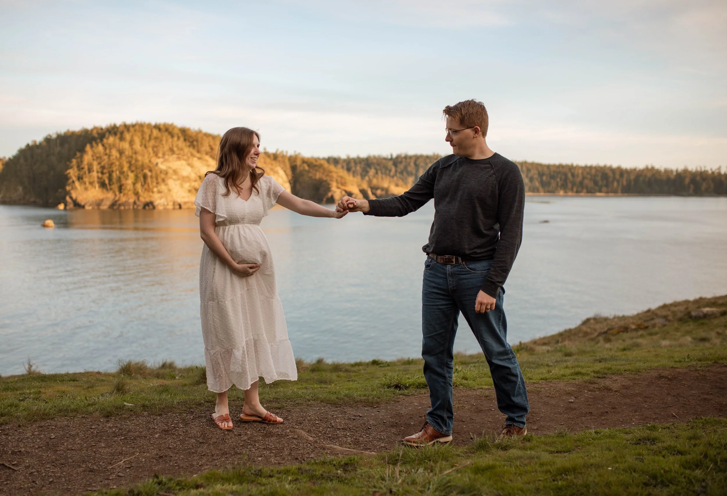 A pregnant woman in a white dress holding her belly and a man in a dark sweater holding her hand by a body of water with a forested hill in the background.