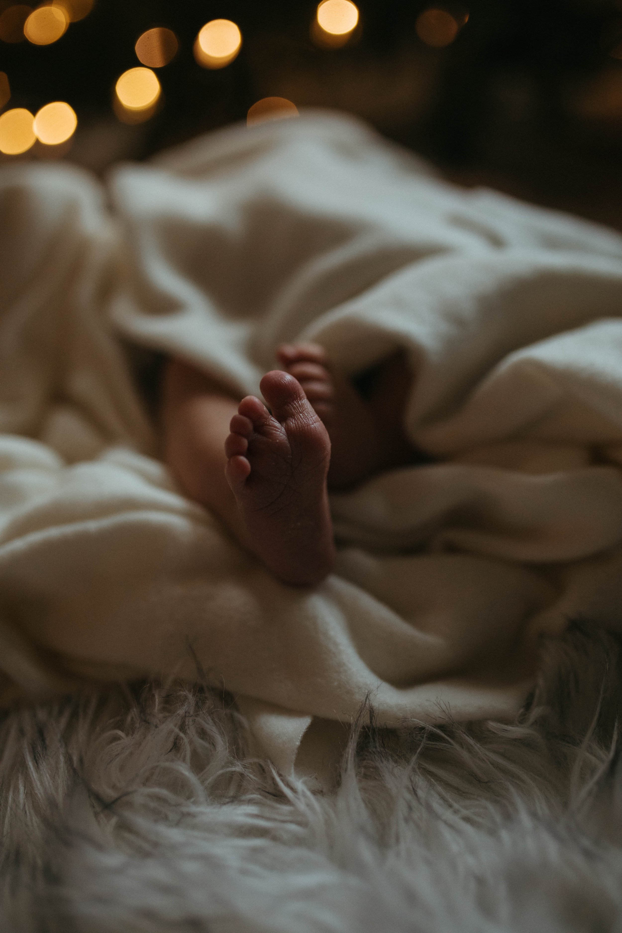 Close-up of a newborn baby's feet, wrapped in white blanket, resting on a soft, furry surface, with warm, blurred string lights in the background.