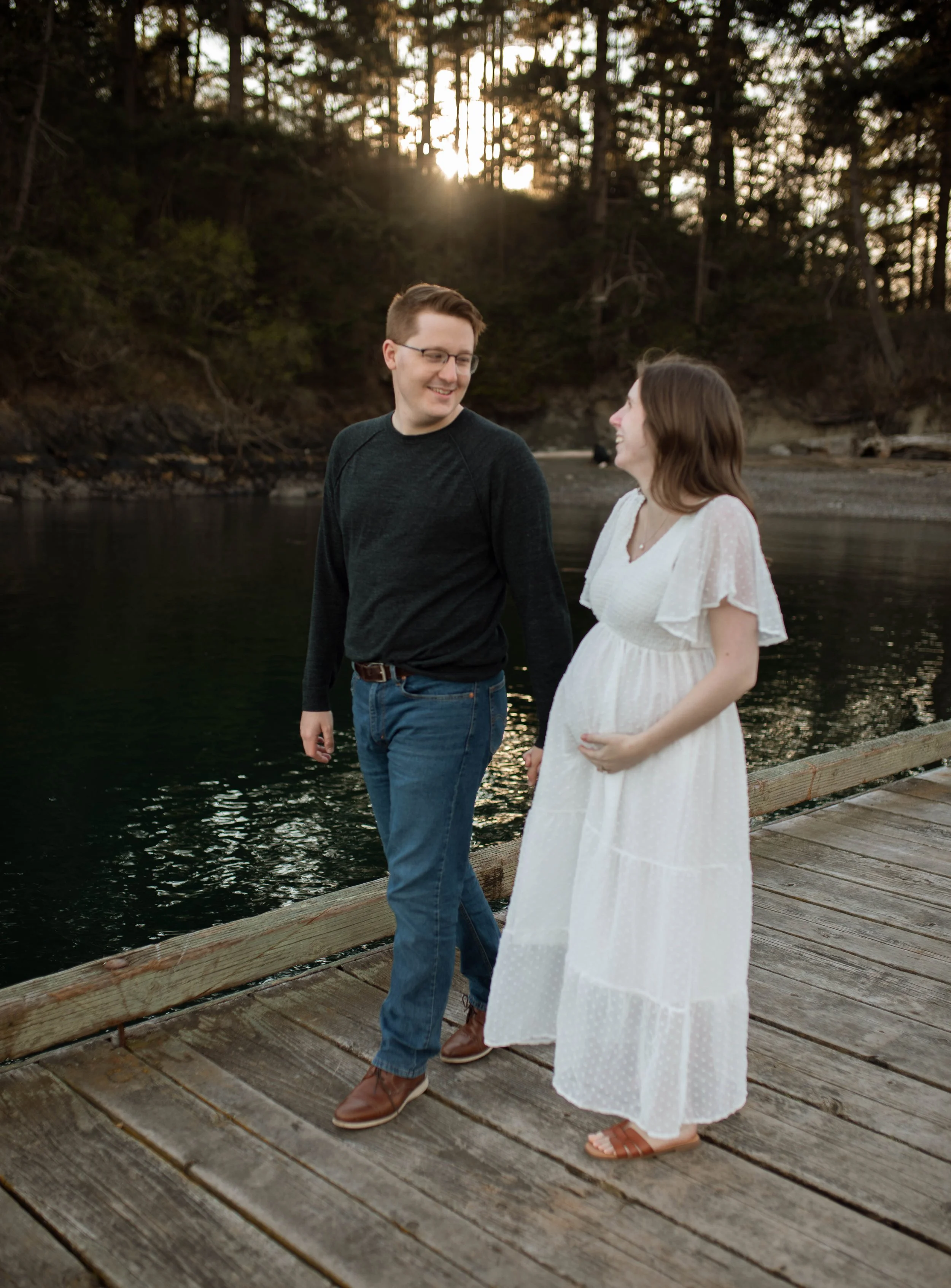 A man and pregnant woman holding hands and smiling on a wooden dock by the water at sunset, surrounded by trees.