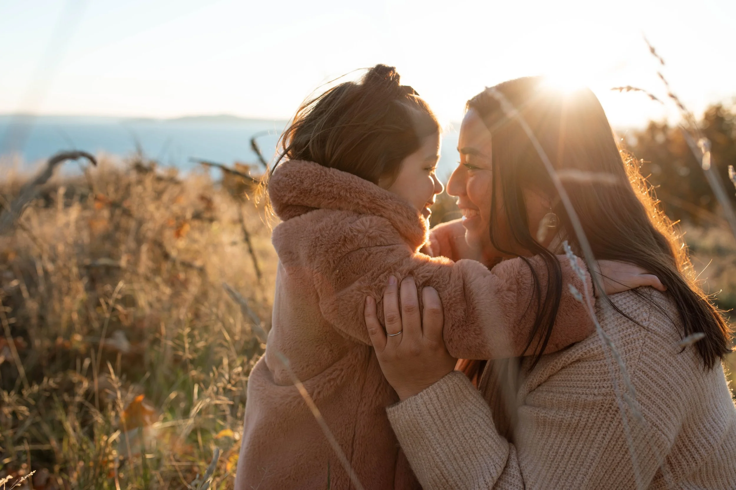 A woman and a young girl share a hug outdoors during sunset, smiling with their faces close together, surrounded by tall grasses and a clear sky.