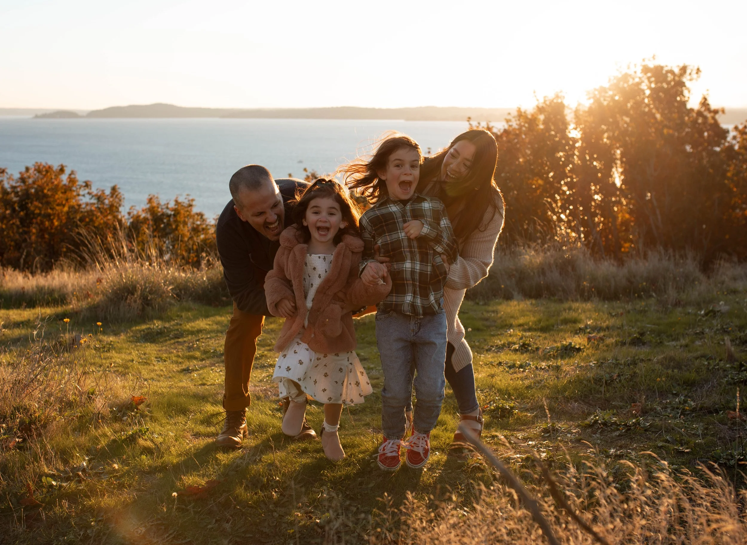 A family of four, including two children, enjoying a sunny outdoor moment near a body of water with trees in the background, during sunset.