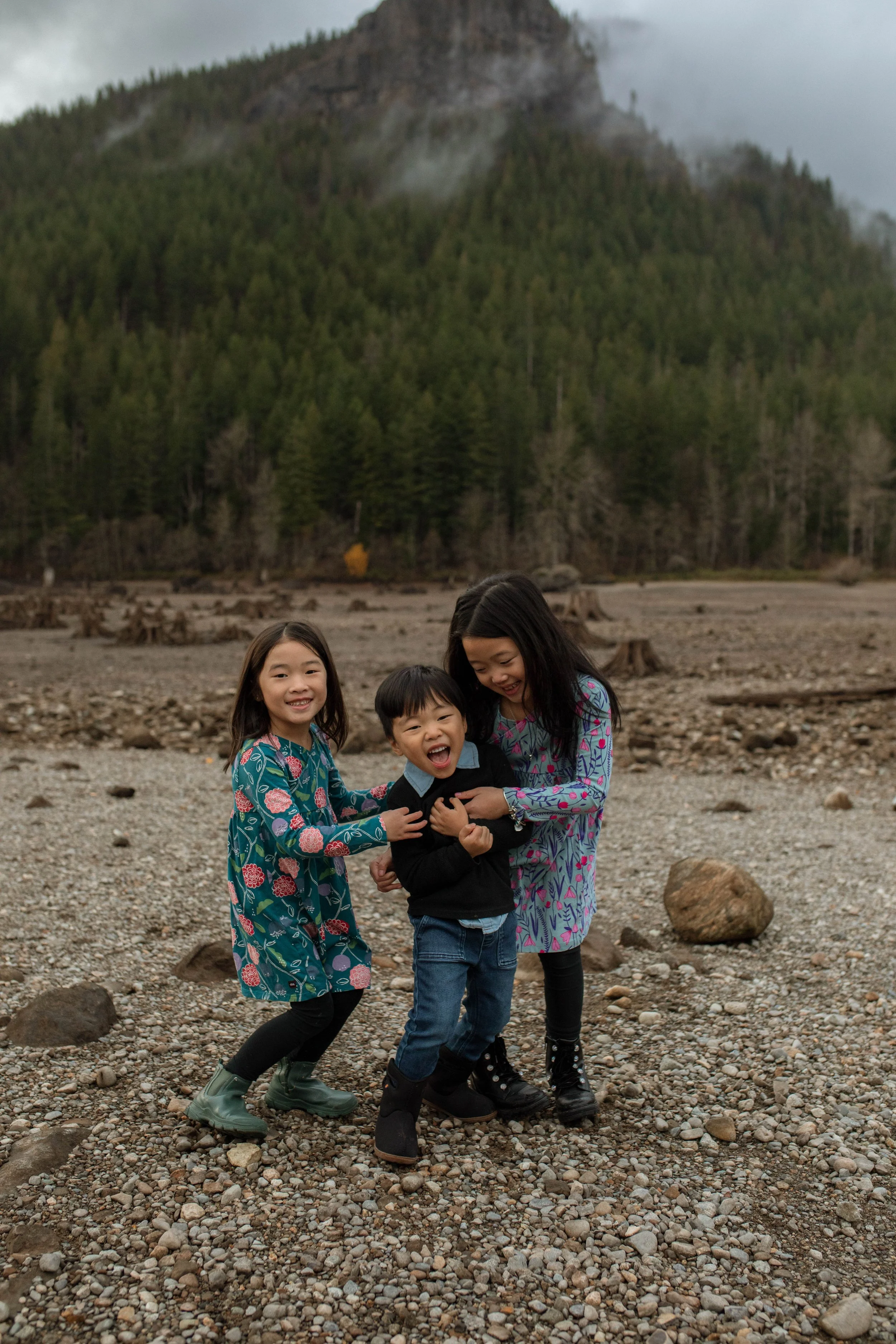 Three children, two girls and one boy, playing and smiling on a rocky outdoor terrain with a mountainous forested background.