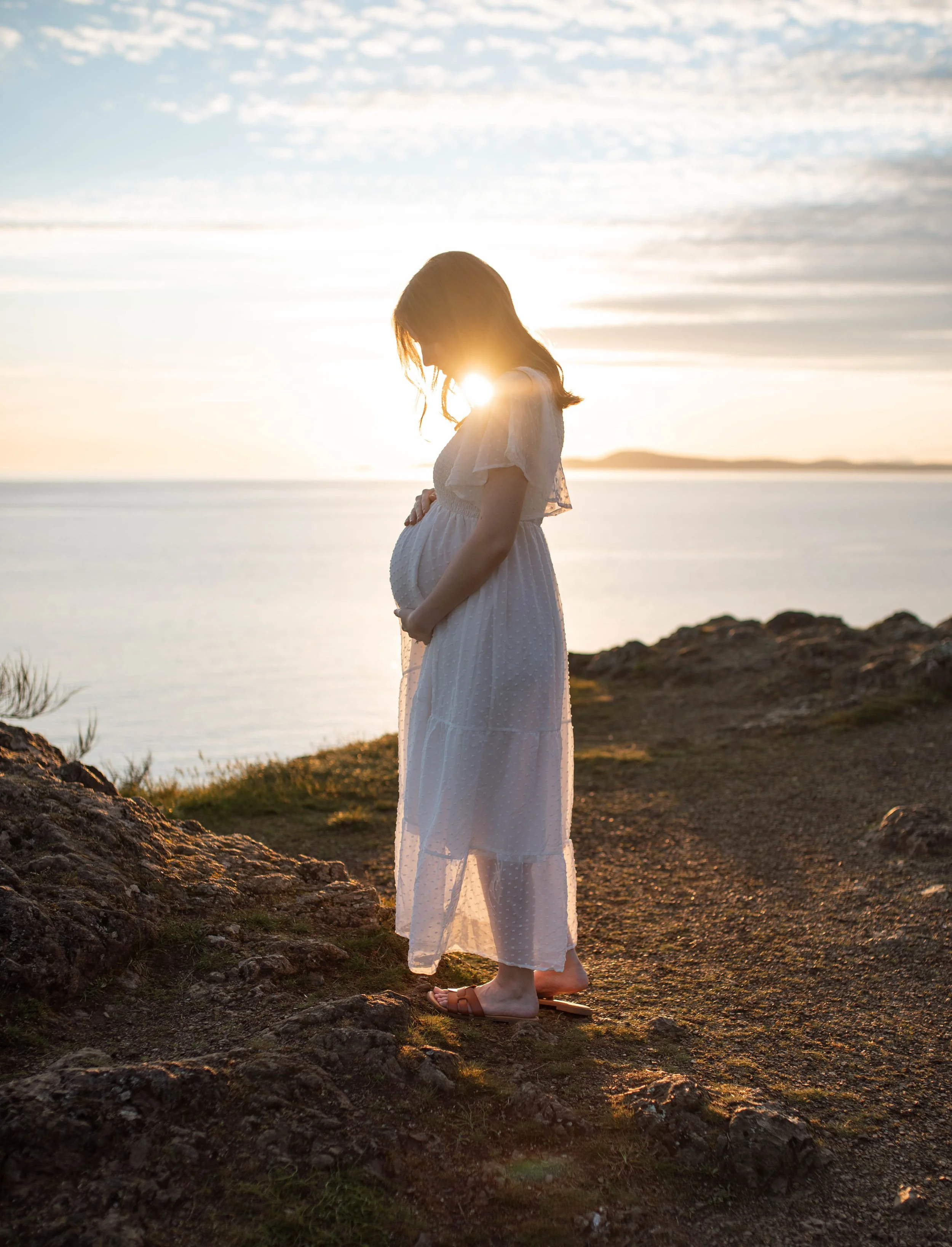 Pregnant woman in white dress standing on rocky terrain near water during sunset, with the sun behind her.
