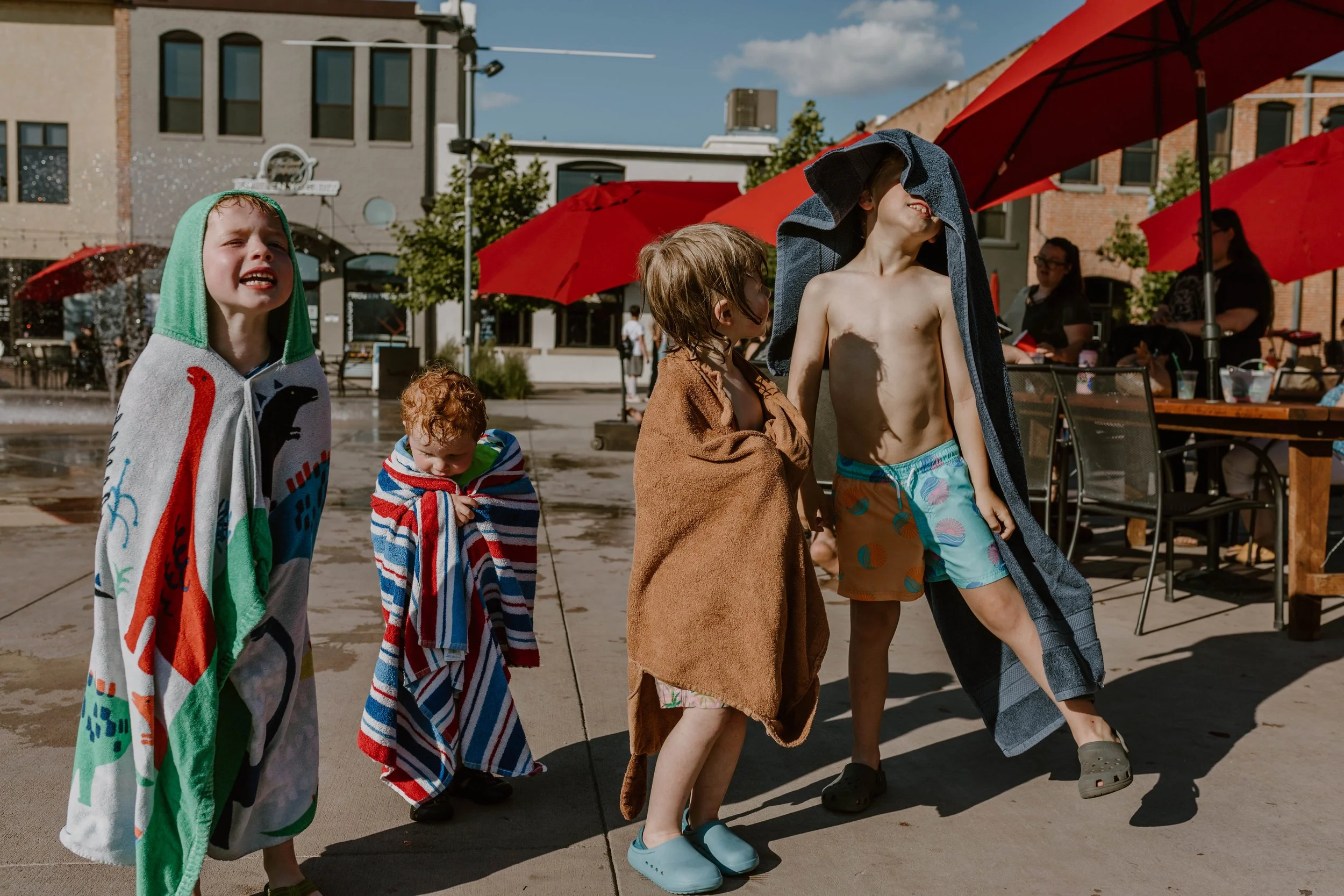 Group of children in swimsuits and towels at a water park, with water spraying and red umbrellas in the background.