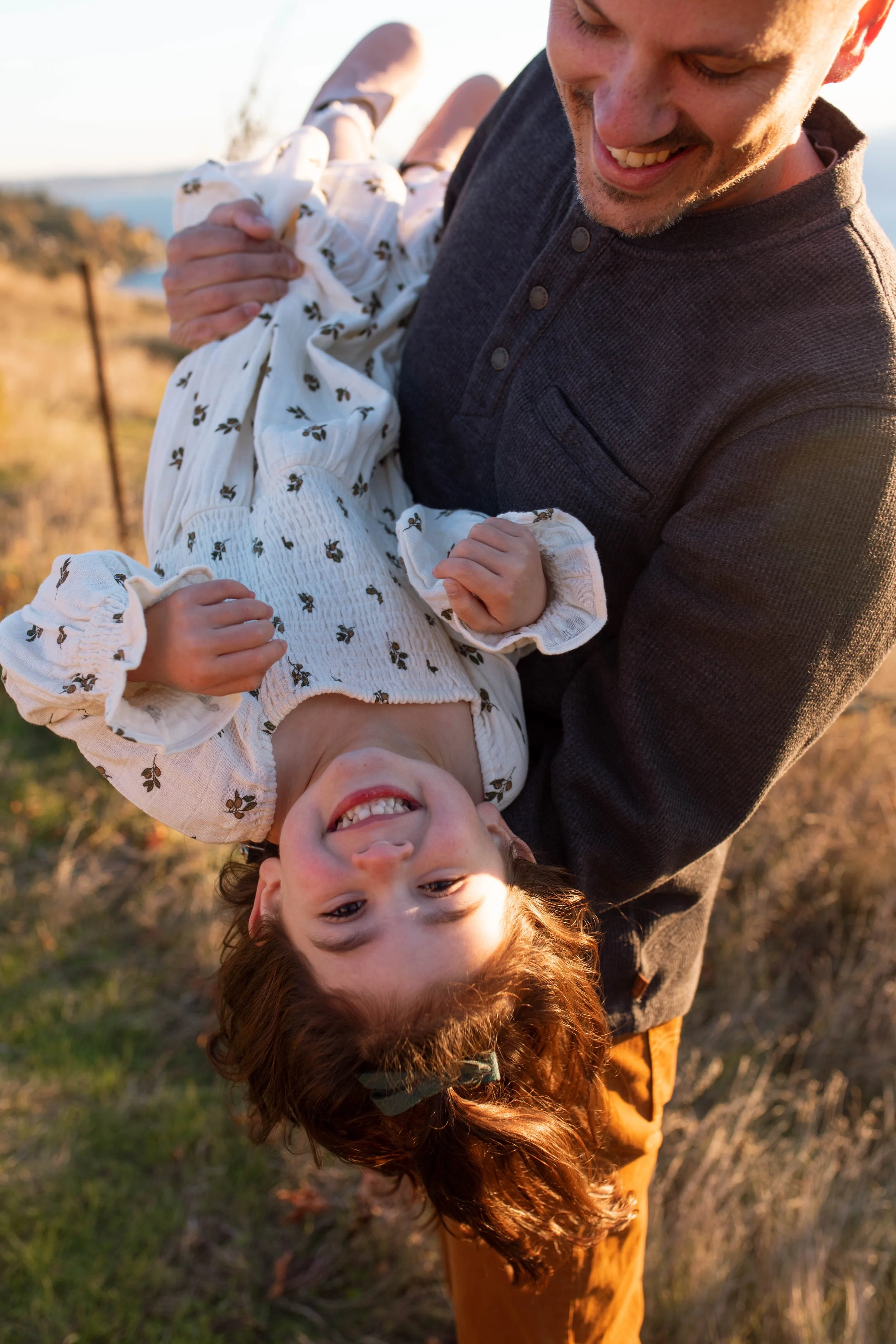 A man holding a smiling young girl upside down outdoors during sunset.