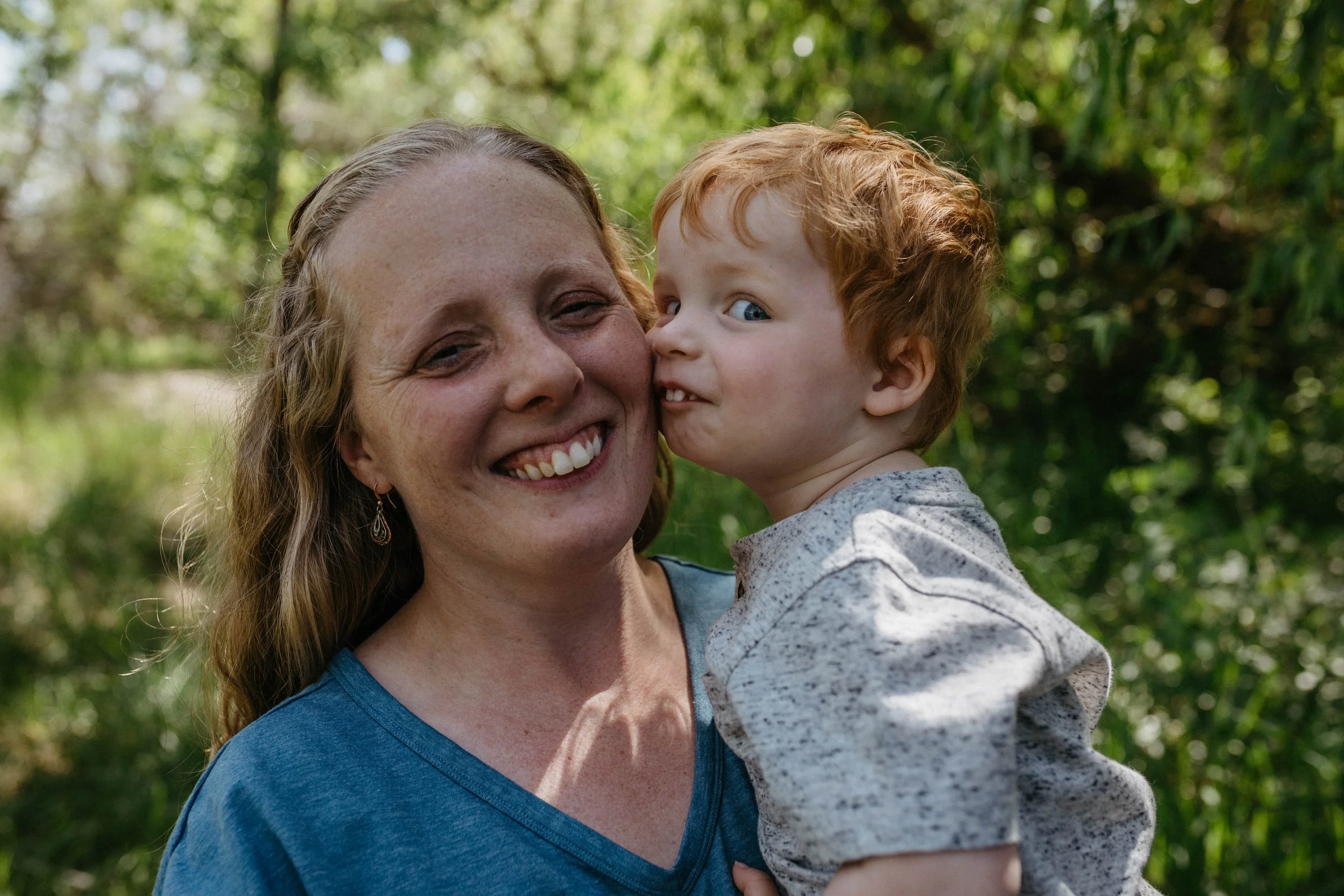 A woman smiling while holding a young boy with red hair and blue eyes, outdoors with green foliage in the background.