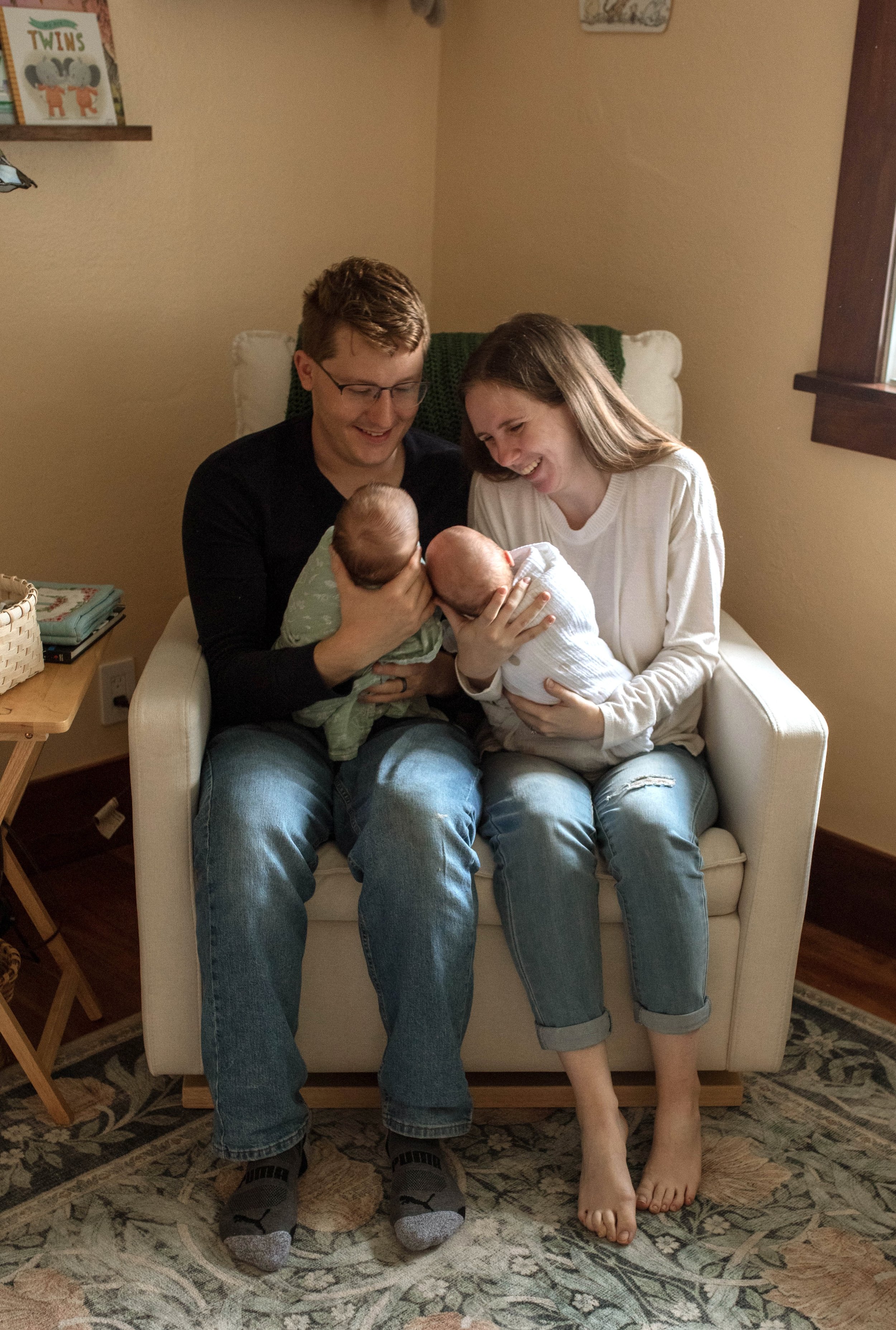 A family of four sitting on a beige armchair in a cozy home. The father and mother, both smiling, are holding twin newborn babies. The father is wearing glasses, a black shirt, and jeans, while the mother is in a white long-sleeve shirt and ripped je